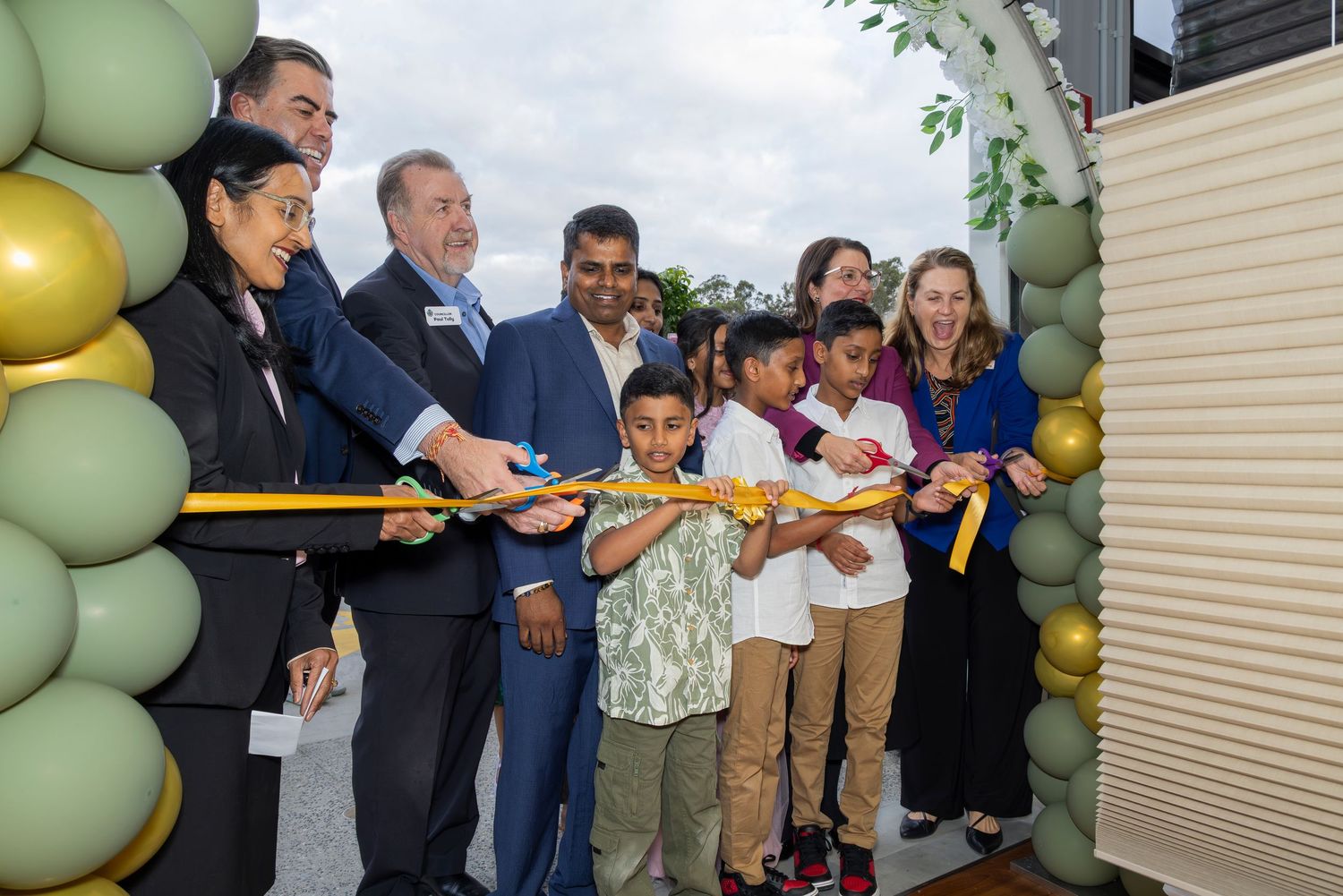 Group ribbon-cutting ceremony with adults and children under balloon arch at grand opening event.