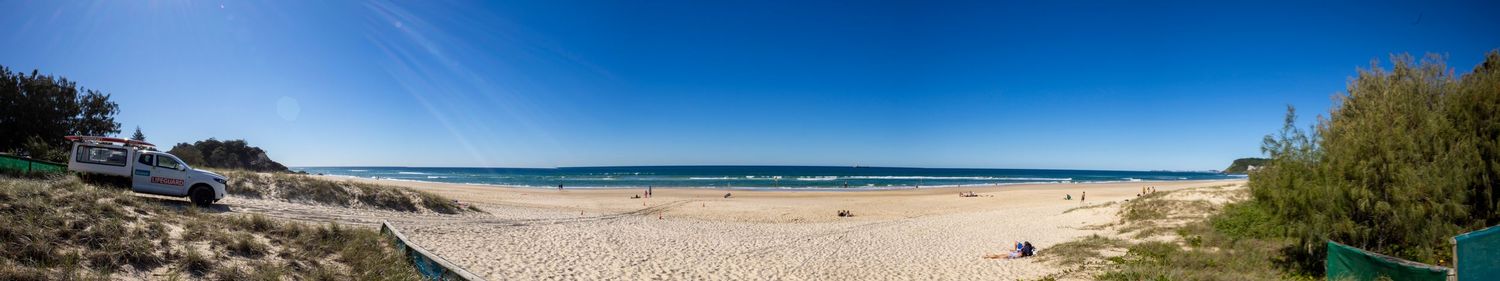 Panoramic view of a pristine sandy beach with clear blue skies, calm waters, and lush green trees lining the shore.