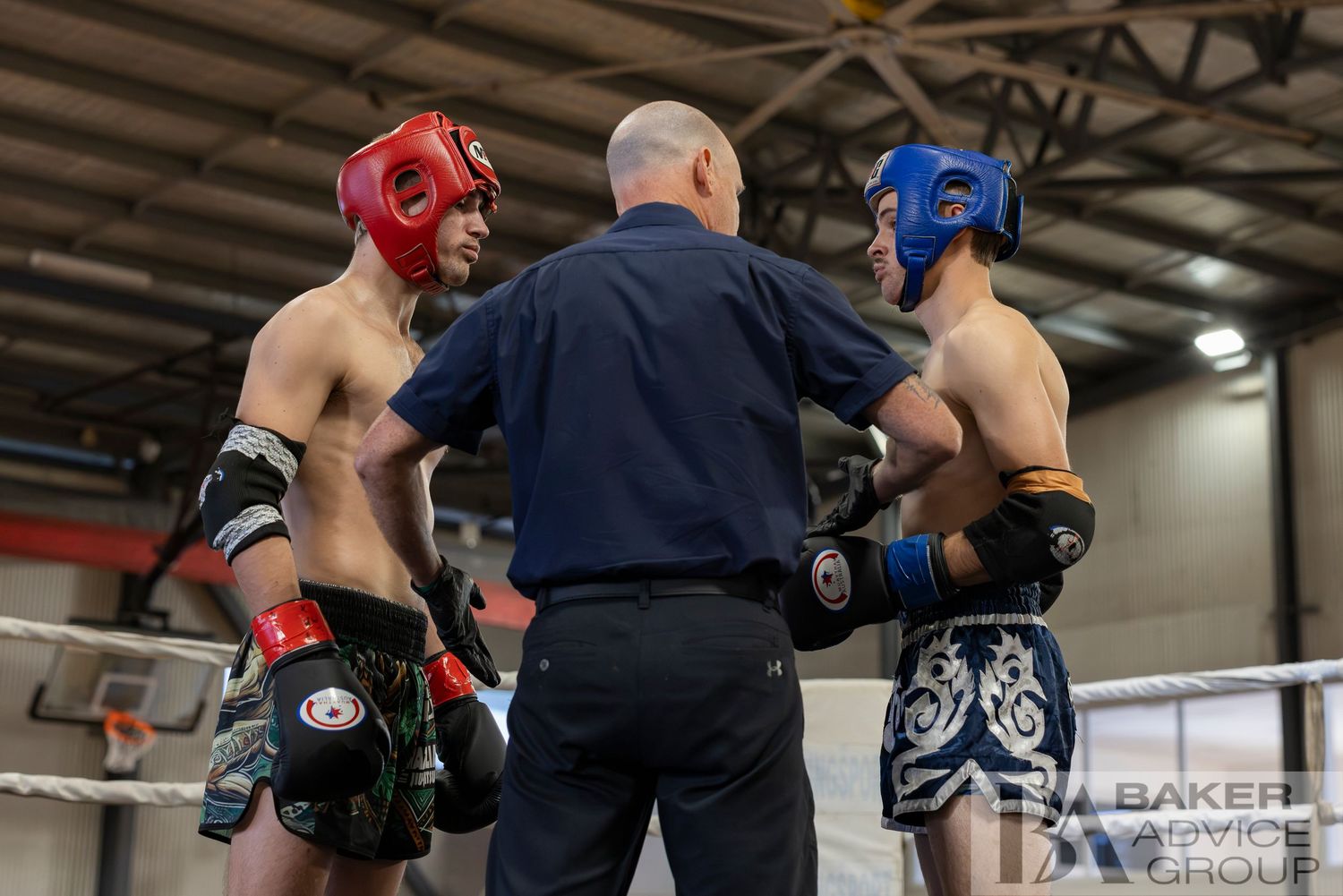 Two young Muay Thai fighters face off in a boxing ring as a referee oversees the bout at an indoor martial arts event.