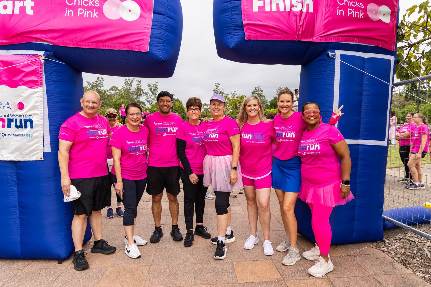 Group of runners in pink shirts posing at Chicks in Pink charity fun run finish line on a sunny day.