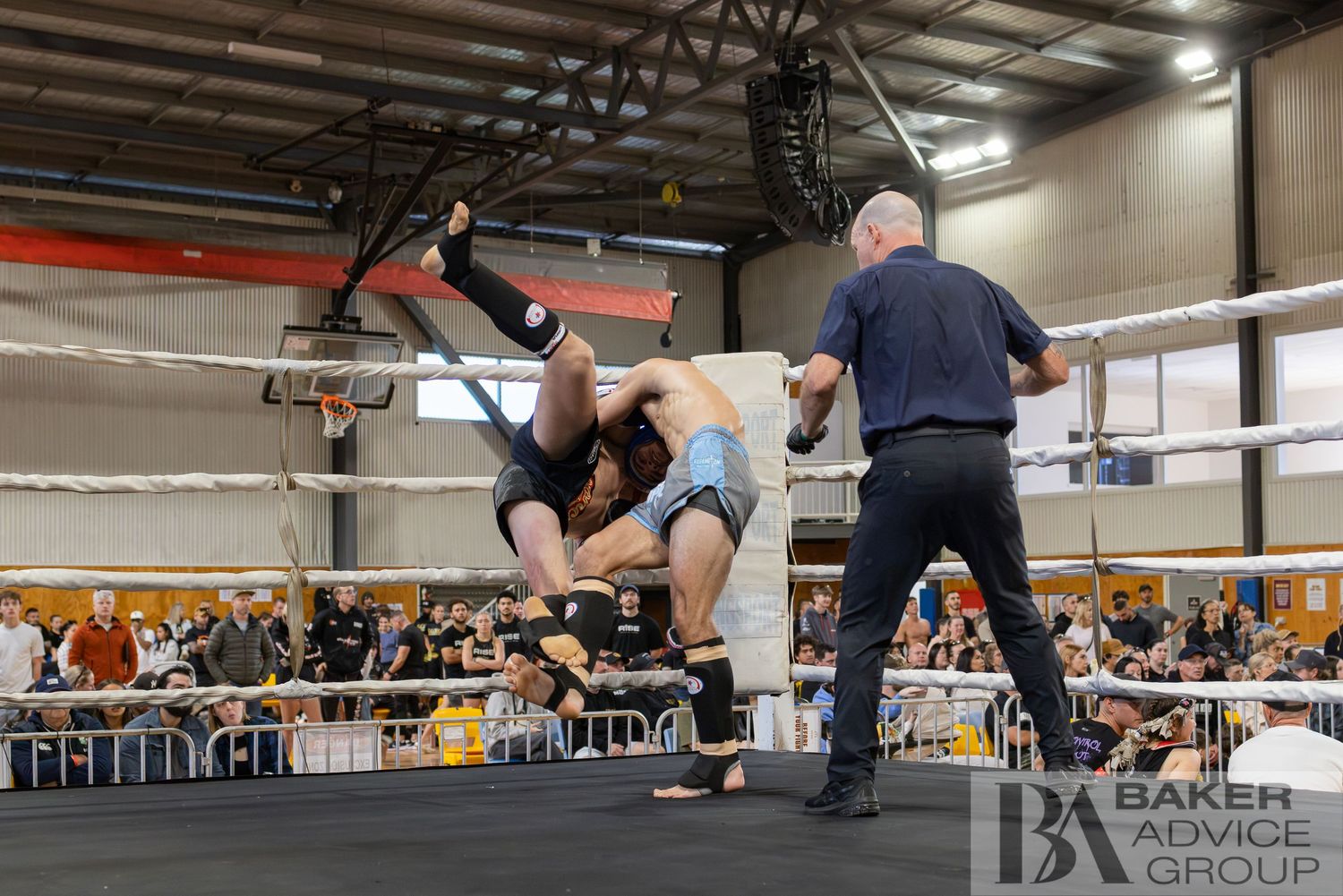 Two wrestlers grapple in a ring as a referee watches during a professional wrestling event indoors.