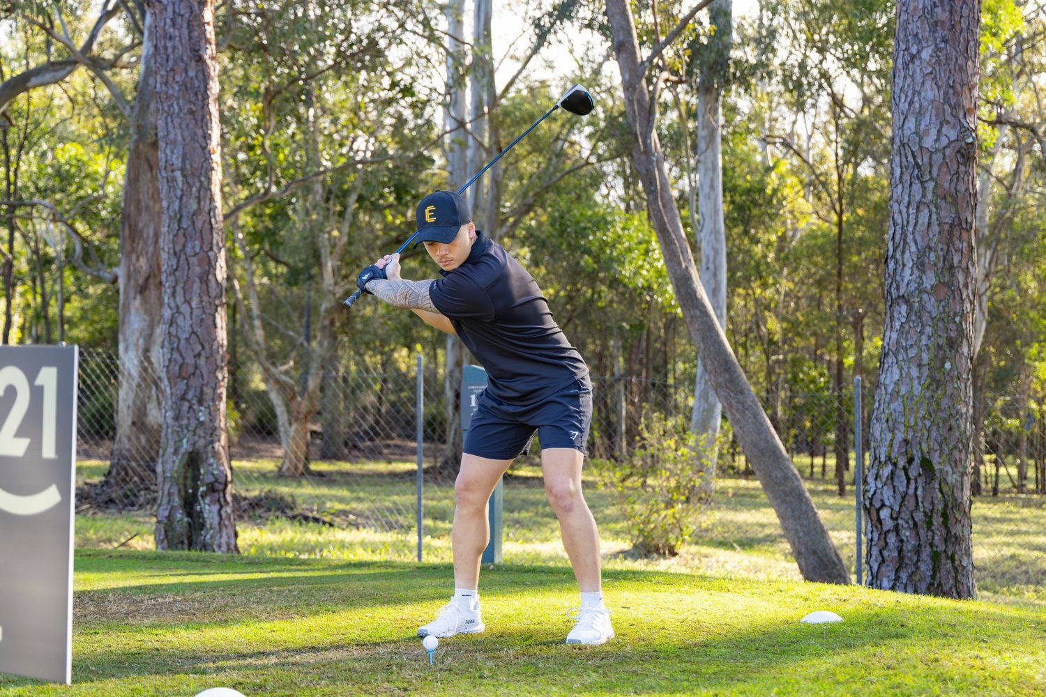 A golfer swings a club at tee box 21 on a tree-lined golf course.