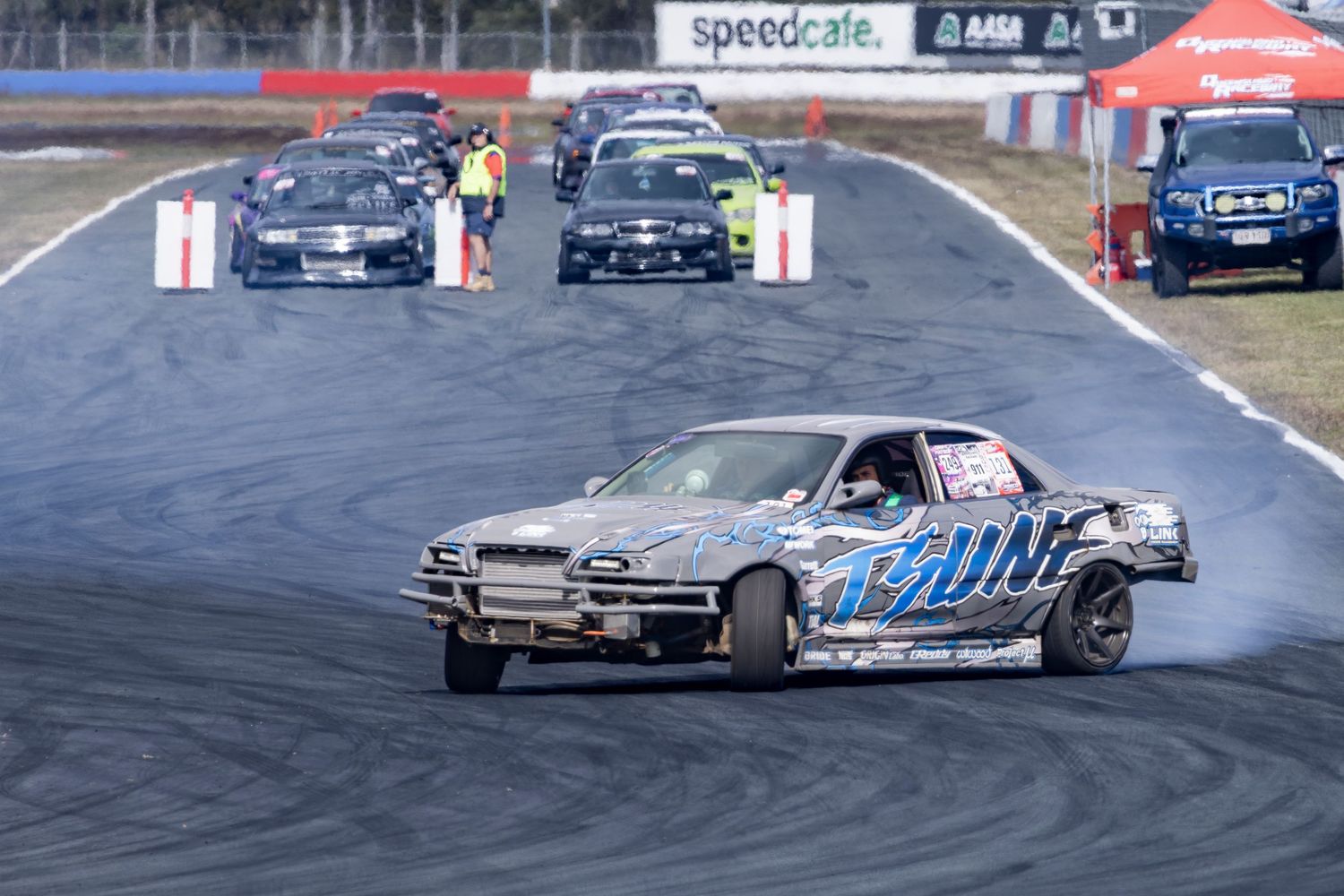 Cars drifting on a race track at a Speedcafe motorsport event, with smoke trailing from tires on a sunny day.