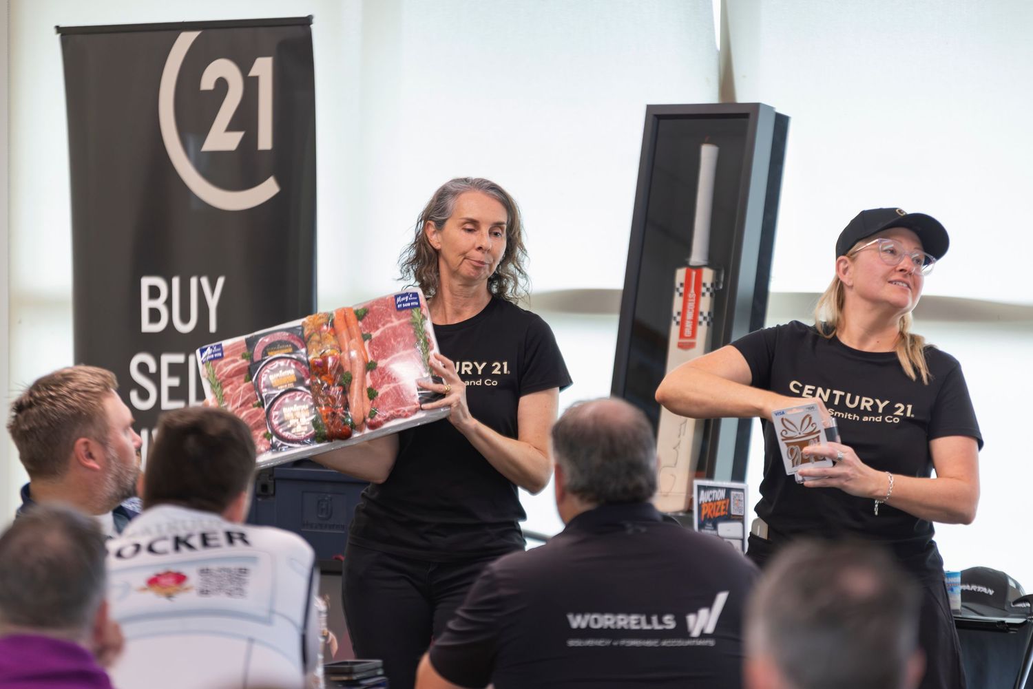 Two Century 21 real estate agents display auction items including a cricket bat and board game at a charity event.
