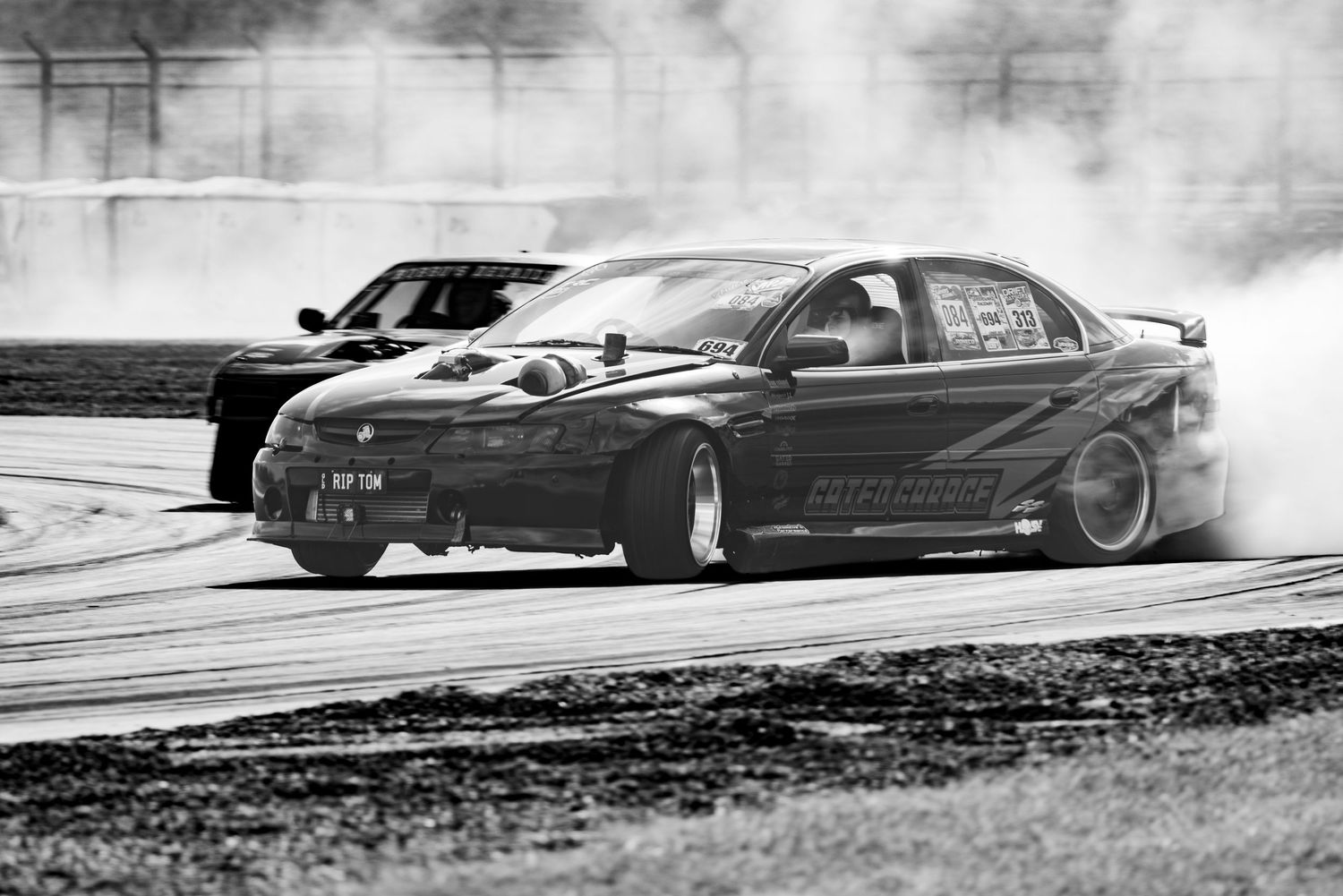 Two drift cars race on a track in black and white, tires smoking as they slide through a turn.