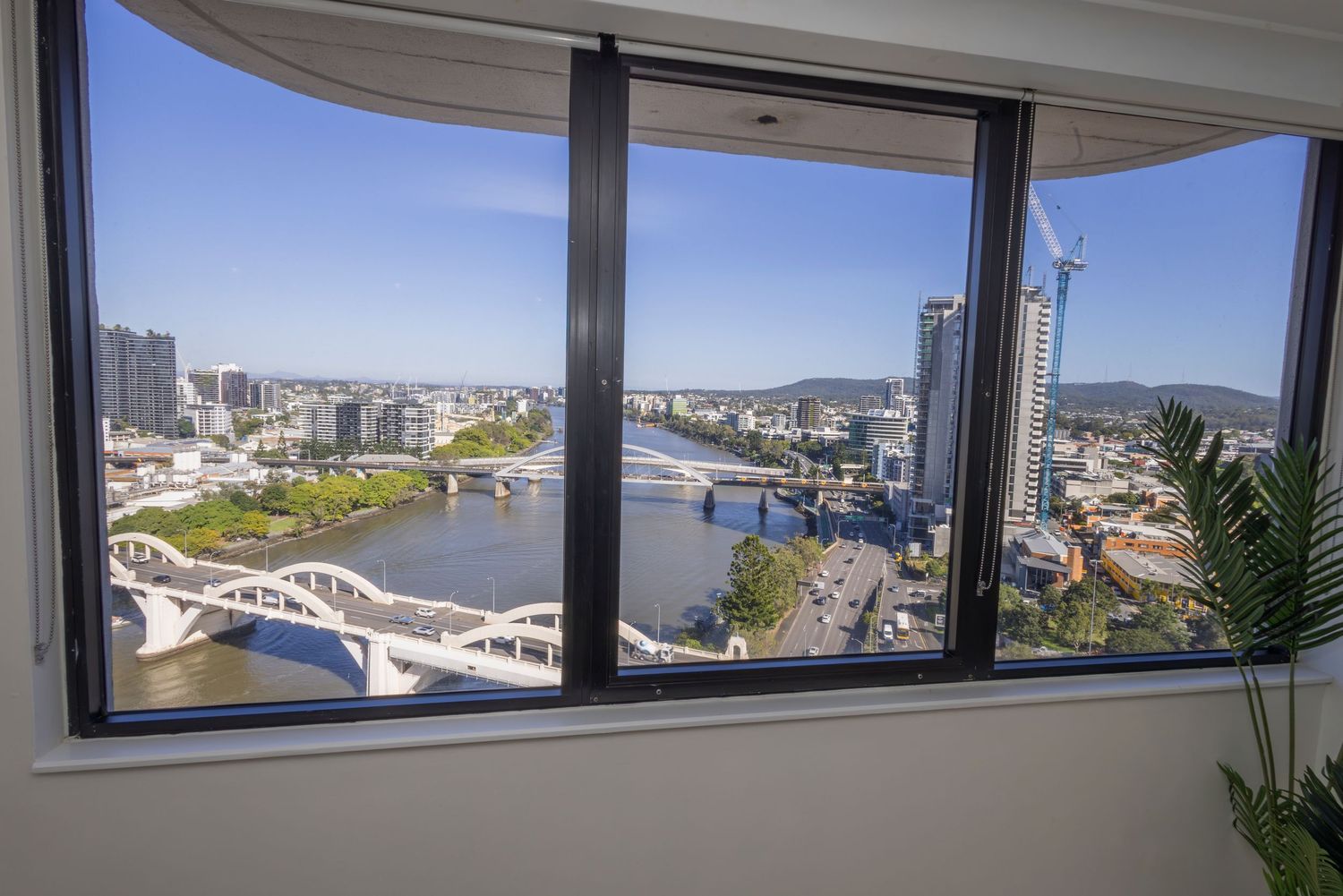 High-rise apartment window view of Brisbane River, bridges, and city skyline on a clear sunny day.