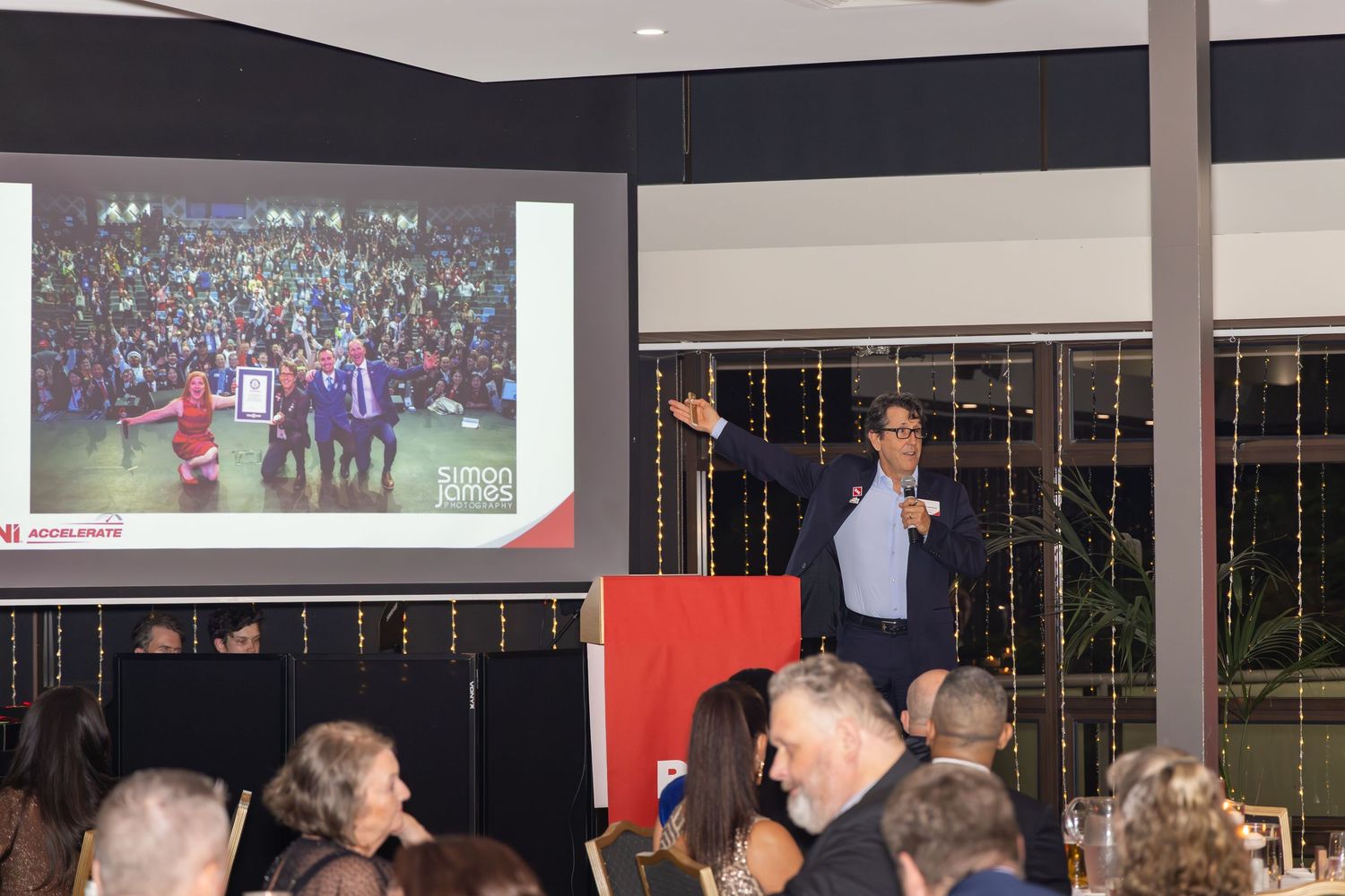 Speaker presents at corporate event with projected crowd image on screen behind him.