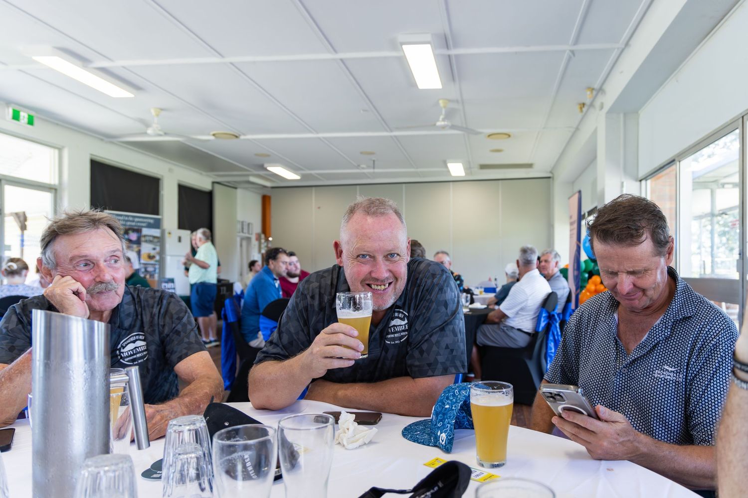 Three men smiling and drinking beer at a golf club social event seated at a table indoors.