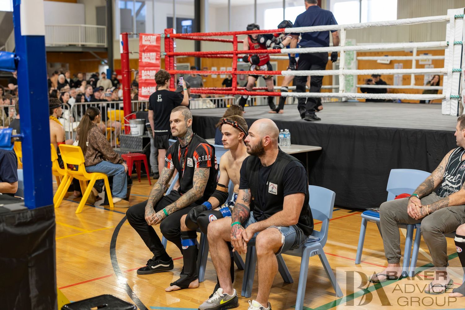 Fighters and coaches rest ringside at an indoor boxing tournament with a competition ring visible in the background.