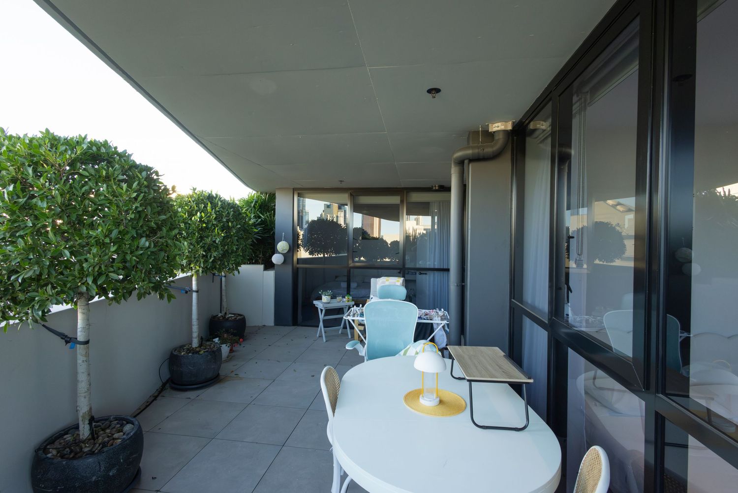 Spacious apartment balcony with outdoor dining set and potted trees overlooking the city.