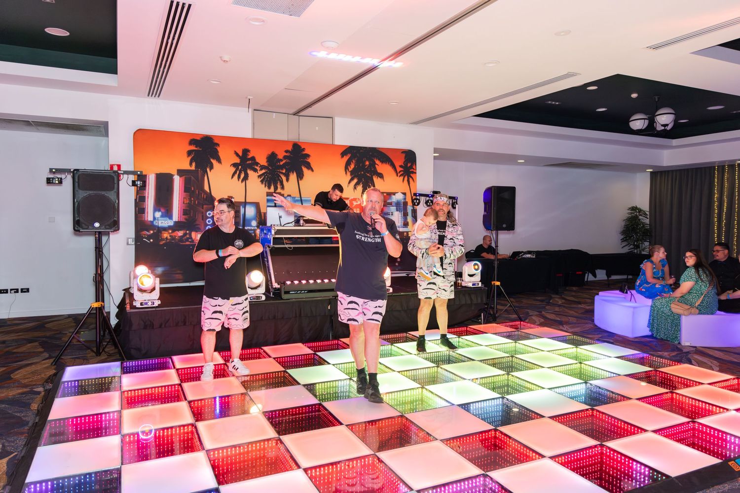 People dancing on a colorful LED dance floor at an indoor party with tropical backdrop and DJ setup.