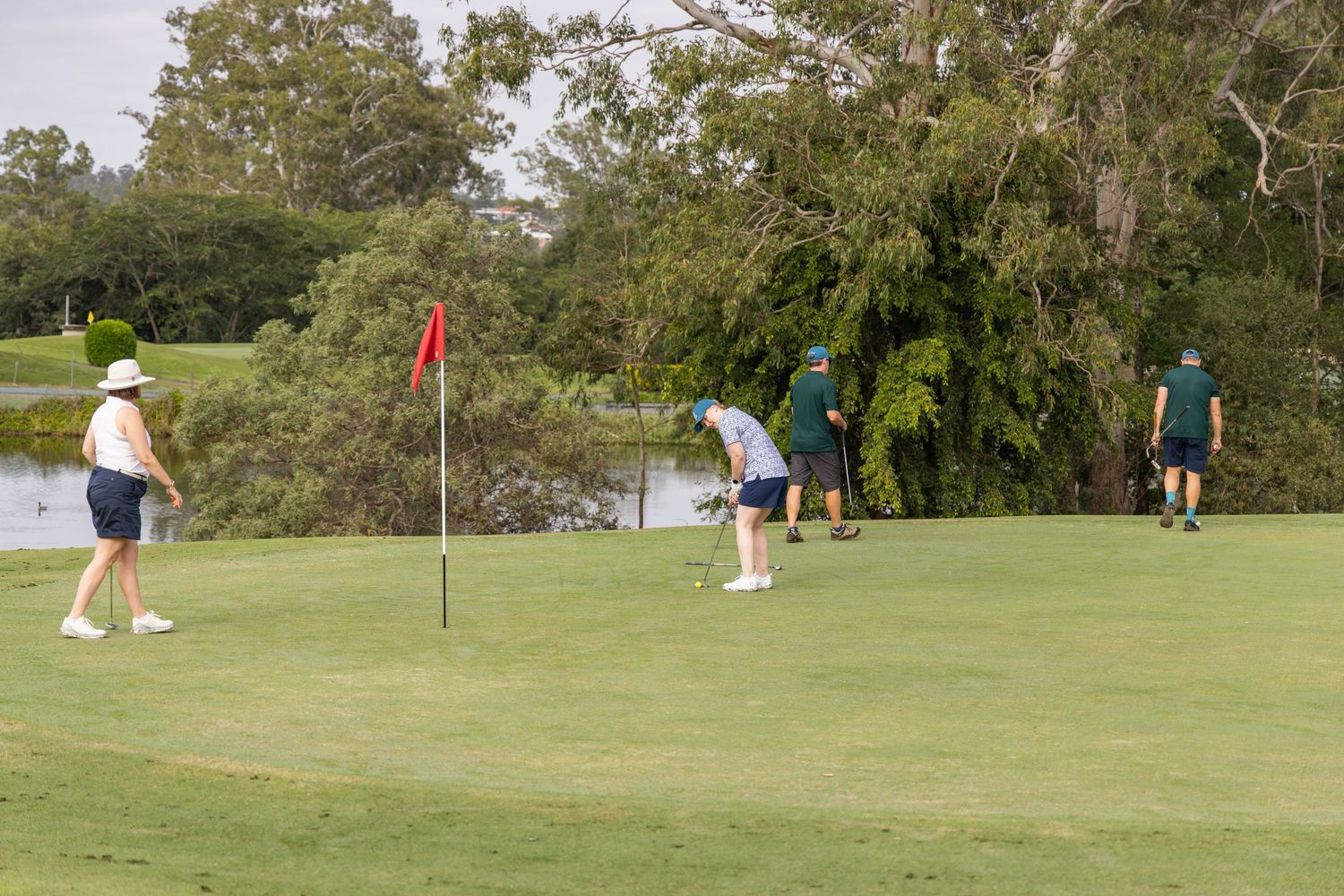 Golfers playing on a lush green golf course near a water hazard, with trees in the background on a sunny day.