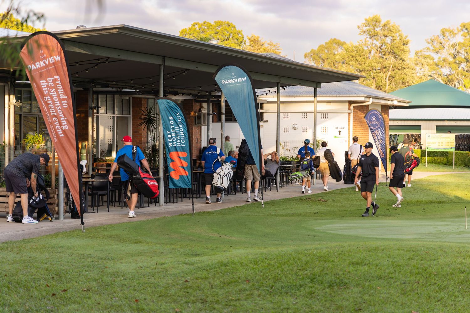 Golfers gather outside a clubhouse with colorful promotional banners on a sunny day at a golf course.