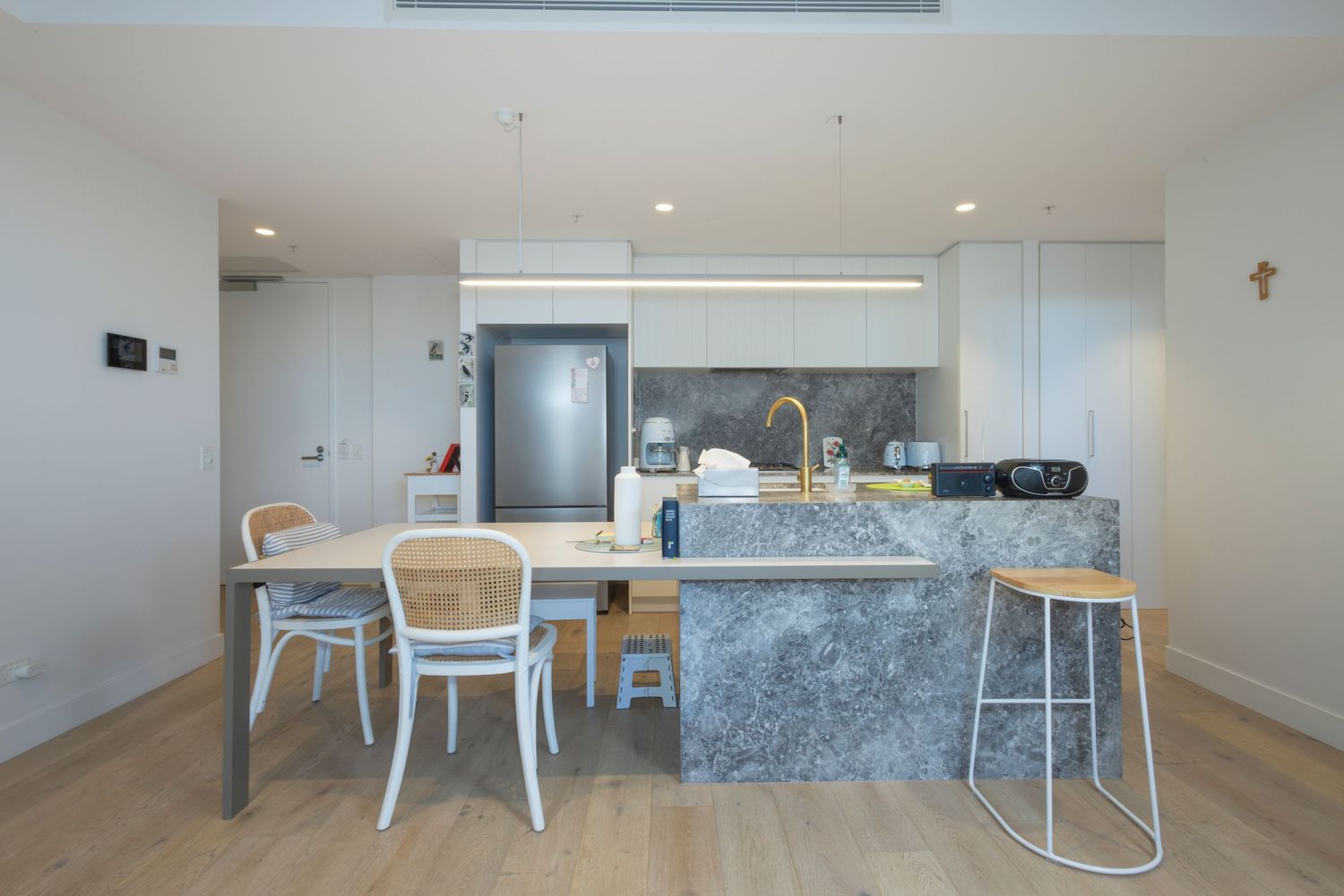 Modern kitchen with marble island, white cabinets, wooden dining table, rattan chairs, and bar stools on light hardwood floors.