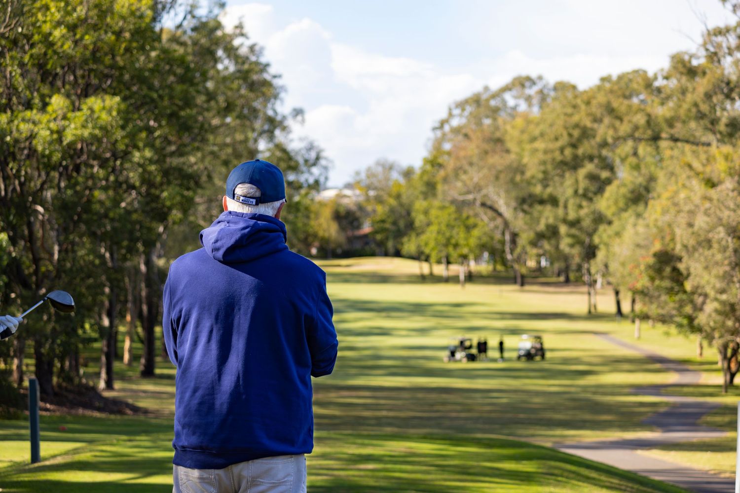 A golfer in a blue hoodie and cap stands on a lush green fairway, surveying the course on a sunny day.
