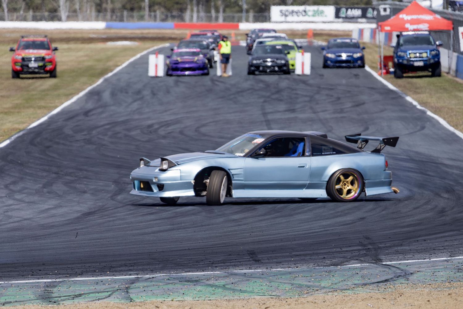 A silver Nissan S13 with gold wheels drifts on a race track, with several cars lined up in the background.