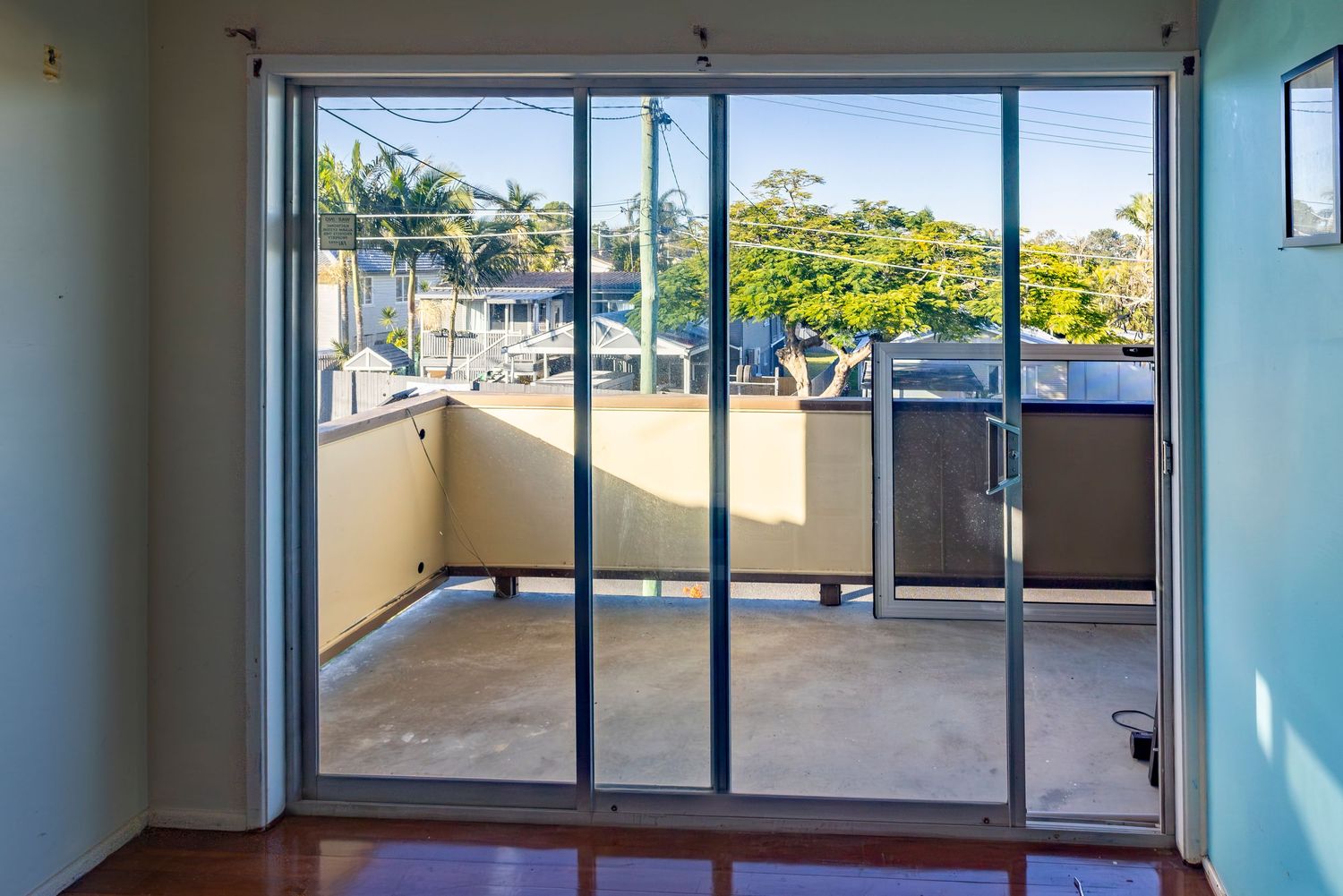Sliding glass doors opening onto a sunny balcony with city and tree views in a modern apartment.