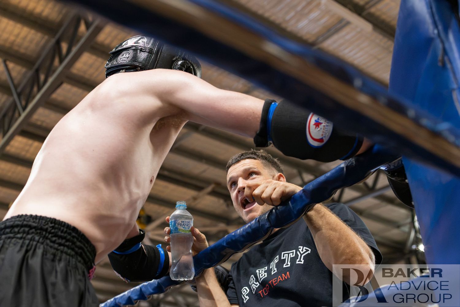 Boxer in headgear throwing a punch in a boxing ring while a coach watches from ringside.
