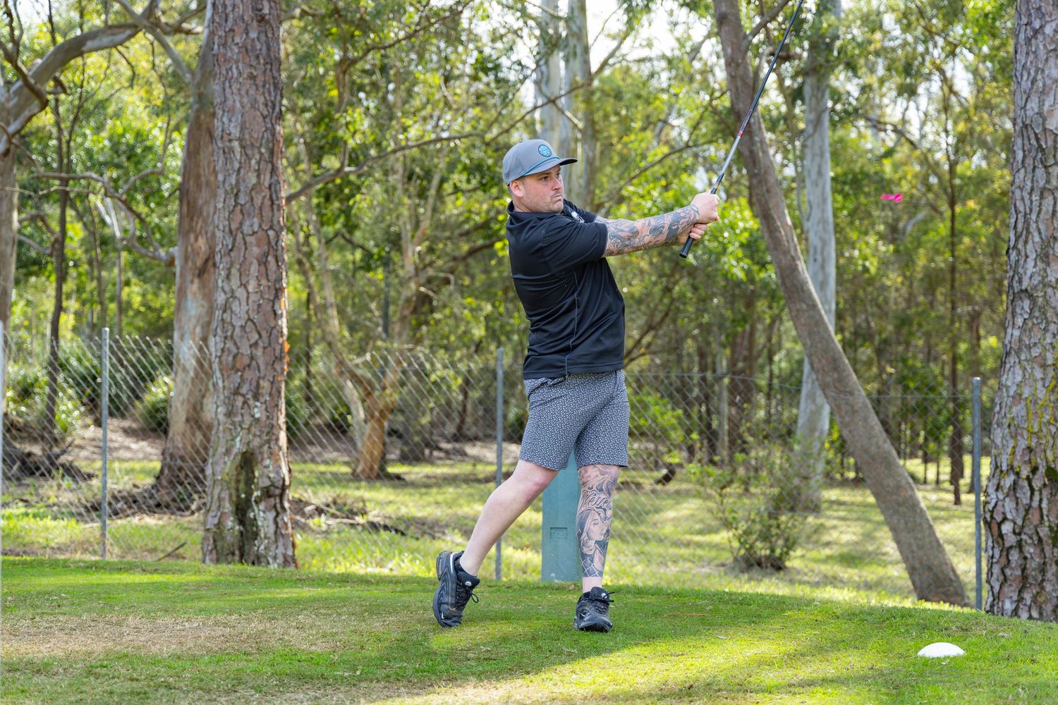 Man playing disc golf in a wooded park, mid-throw stance wearing grey shorts and black shirt.