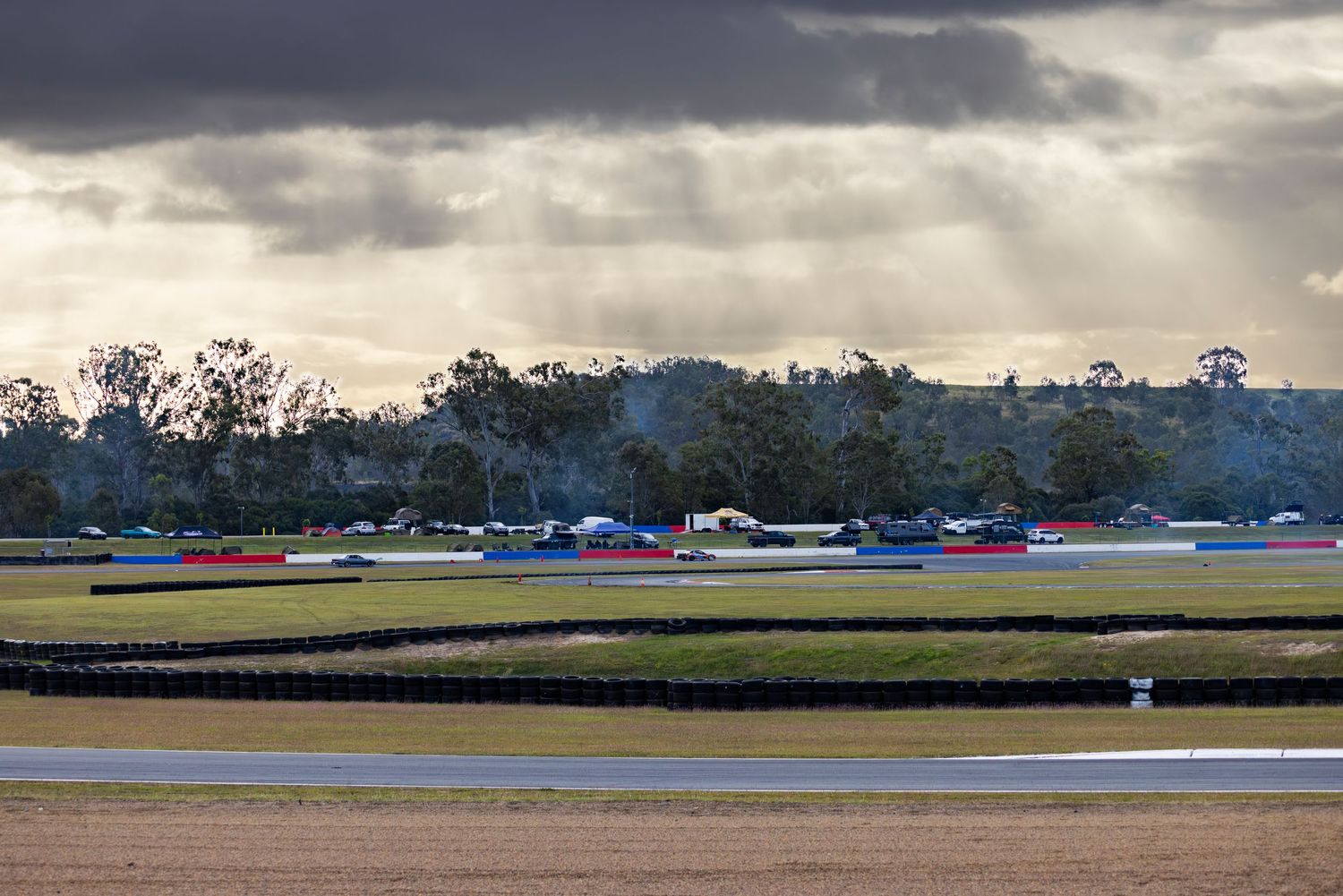 Racing cars speed around a circuit under dramatic cloudy skies with sunrays breaking through at an outdoor motorsport track.