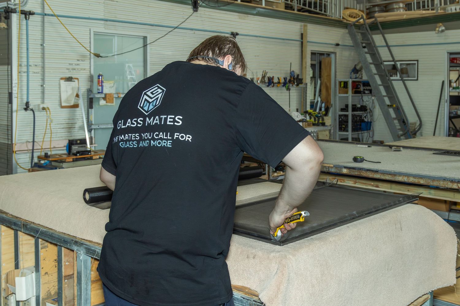 Worker in a black Glass Mates t-shirt cutting material on a workbench in an industrial workshop.
