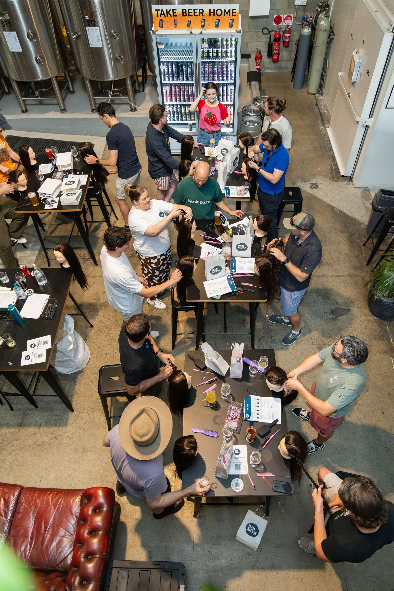 Aerial view of a lively craft brewery taproom with guests gathered around tables sampling beers near a take-home beer fridge.