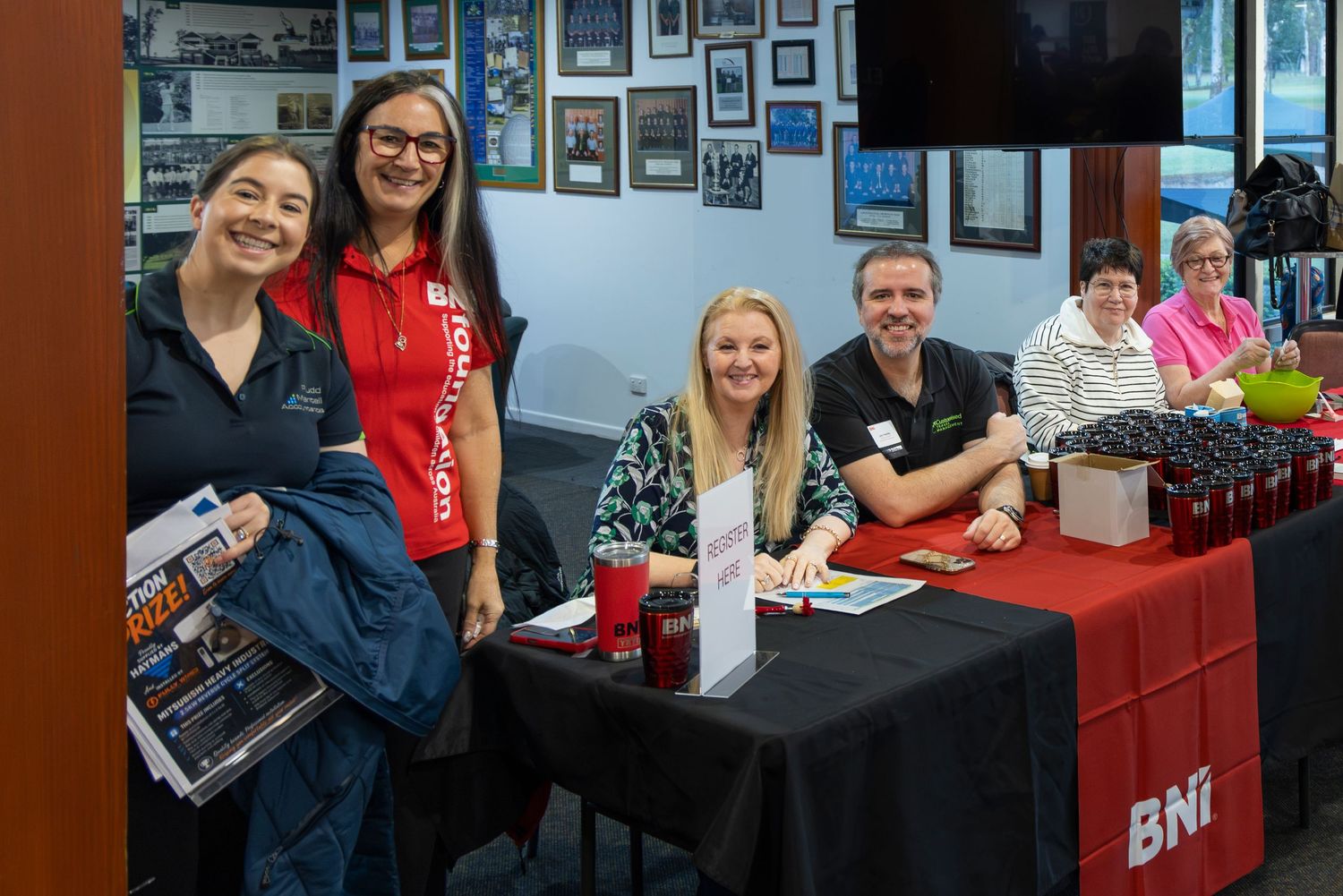 Group of professionals smiling at a BNI networking event table with red cups and promotional materials.