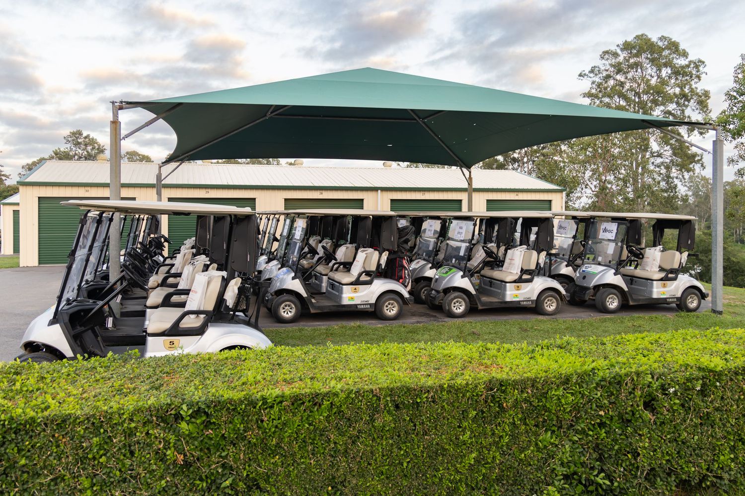 Rows of white golf carts parked under a large green shade structure at a golf course facility.