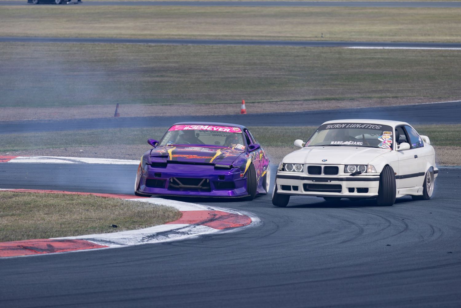 Two drift cars, a purple and a white BMW E36, competing on a race track with tire smoke visible.