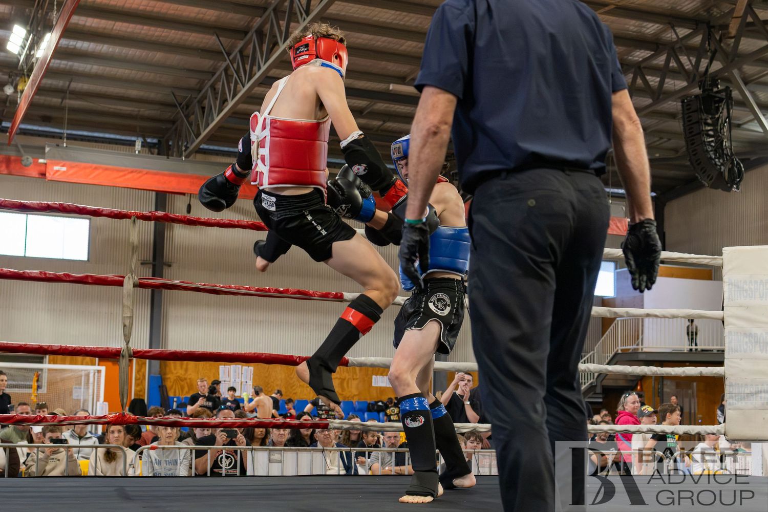 Two kickboxers sparring in a ring during a martial arts competition, with a referee watching and spectators in the background.