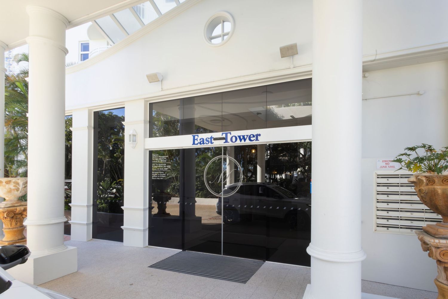 Glass entrance doors of East Tower building with white columns and lush greenery visible in the background.