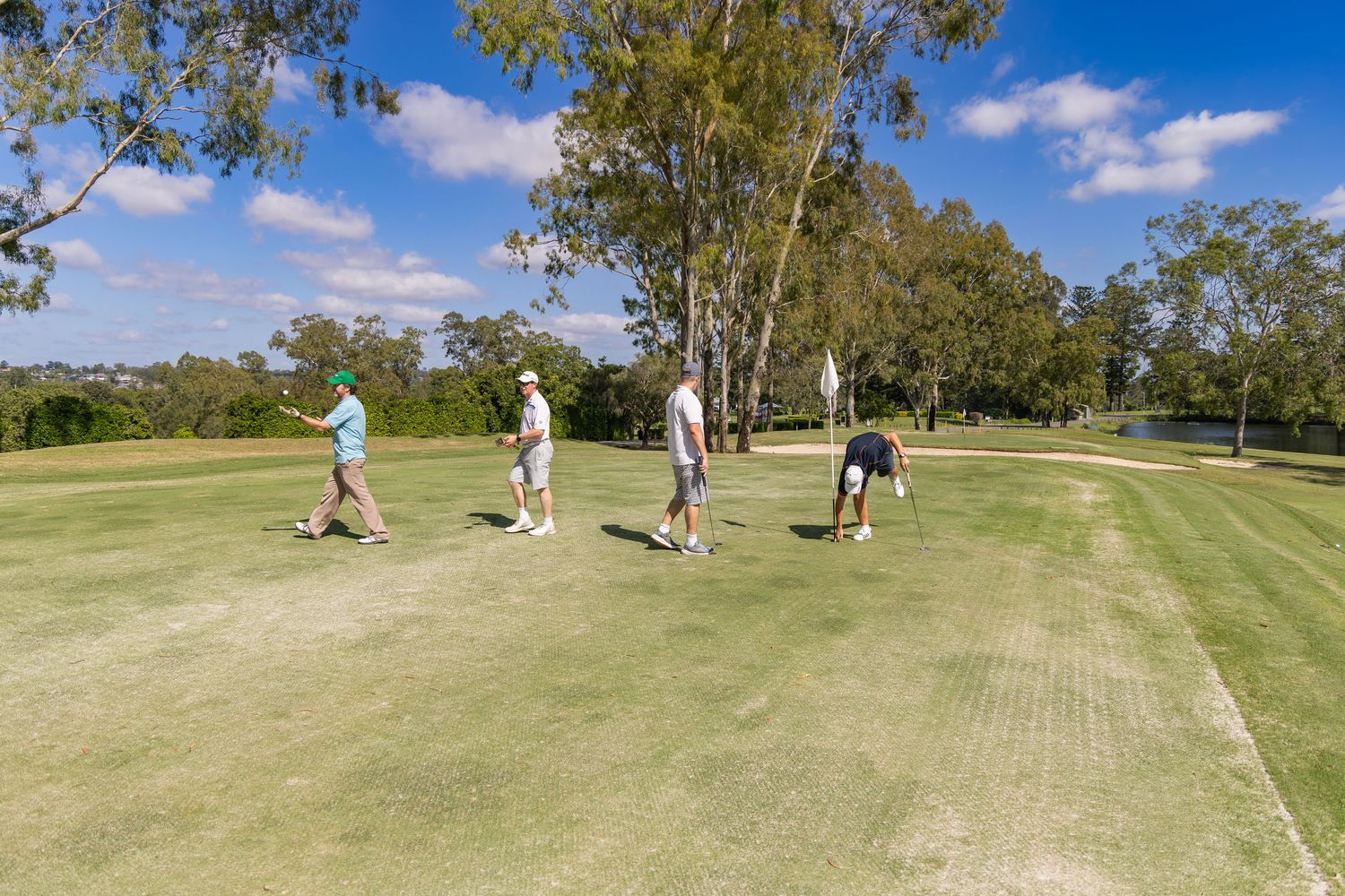 Four golfers playing on a lush green golf course surrounded by tall trees under a bright blue sky.