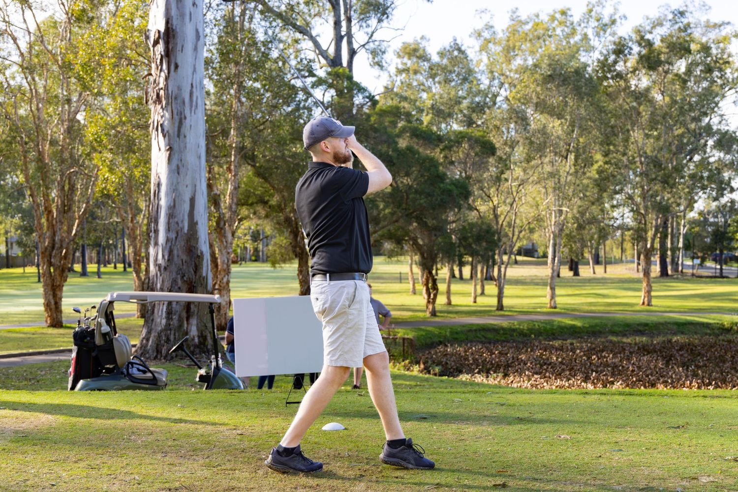 Golfer in black shirt and white shorts completing a golf swing follow-through on a sunny green course.