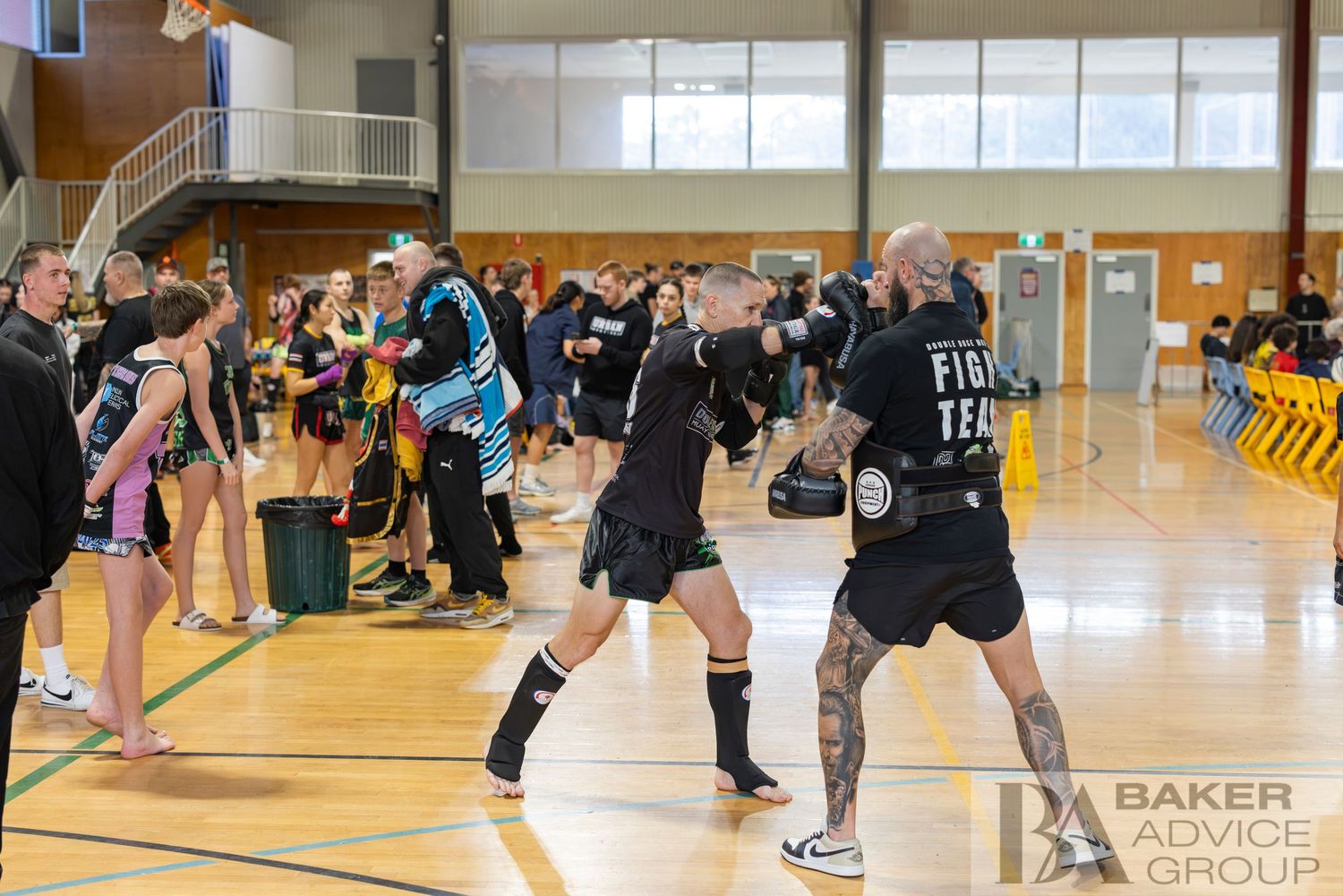 Two fighters spar in a kickboxing training session on a gymnasium floor surrounded by teammates and coaches.