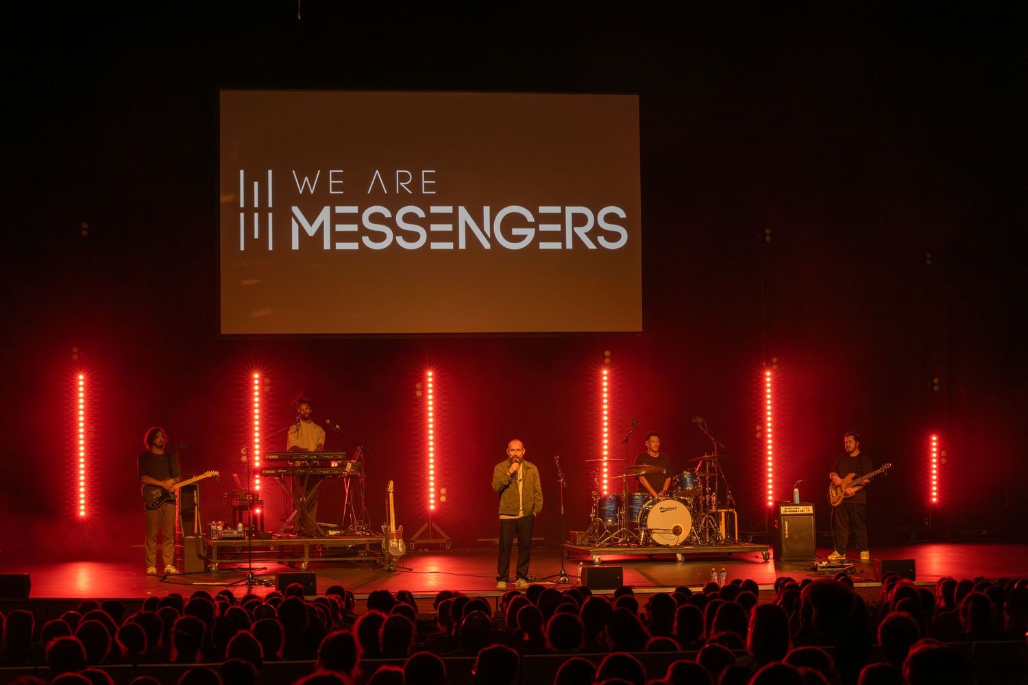 We Are Messengers Christian band performing live on stage with red neon lights and a projection screen displaying their name.