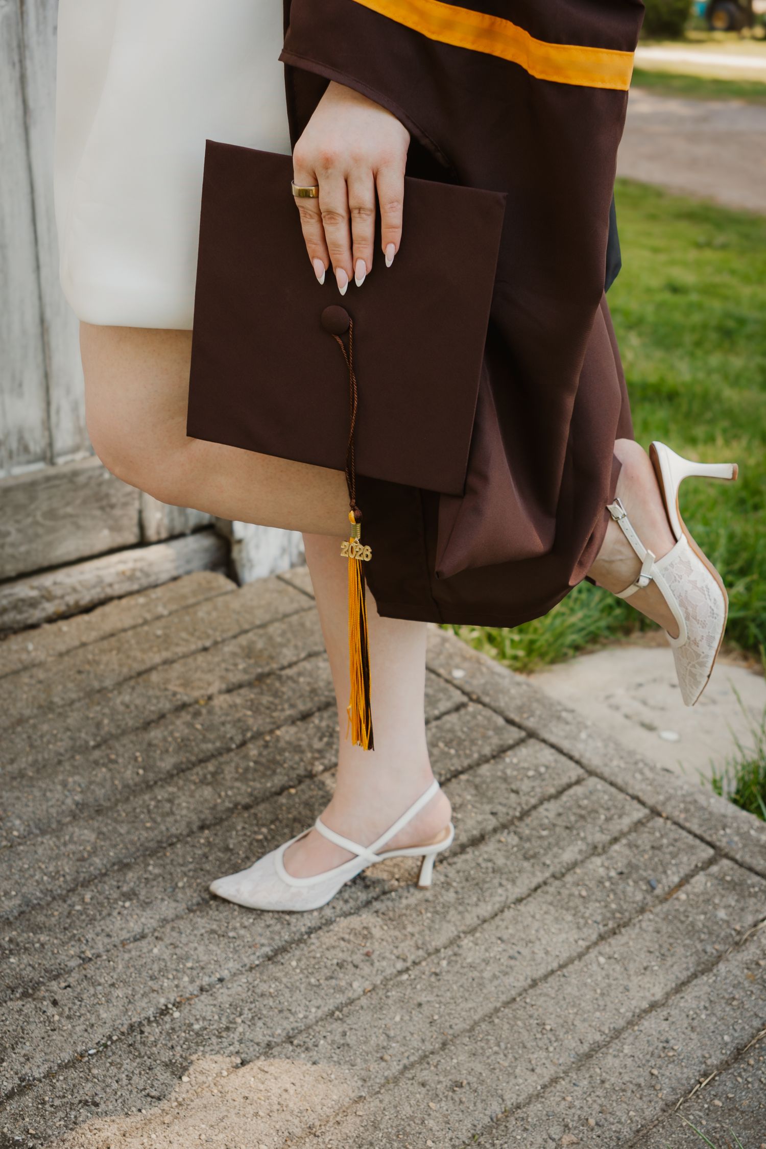 Graduate holding dark maroon cap with gold tassel, wearing white heels and brown graduation gown outdoors.