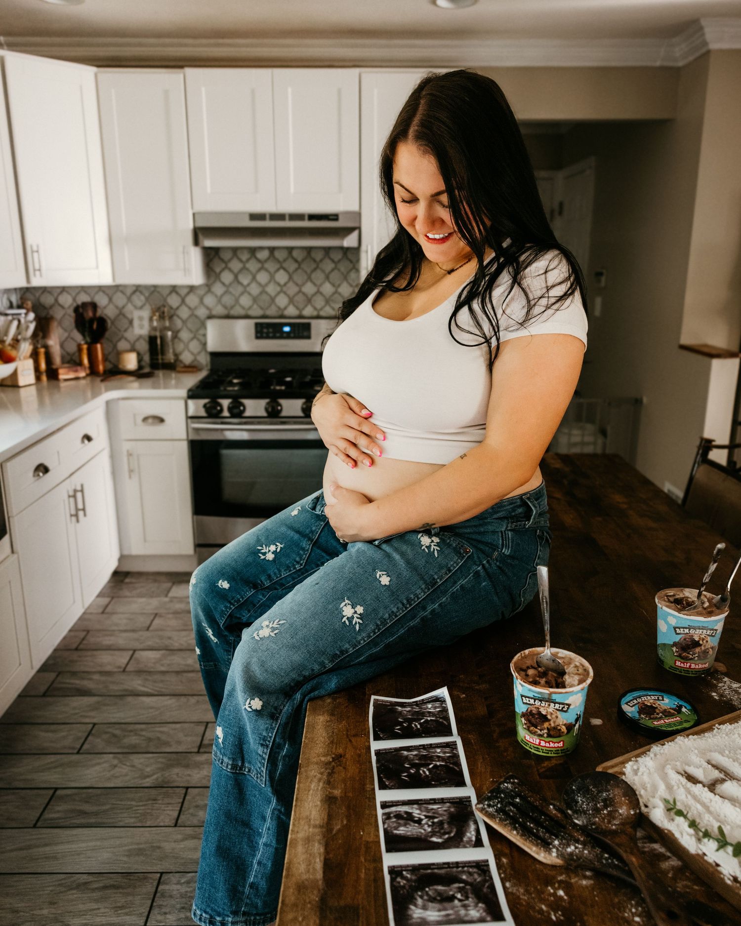 Pregnant woman in white crop top and jeans holds baby bump while sitting on kitchen counter with ultrasound photos and ice cream.