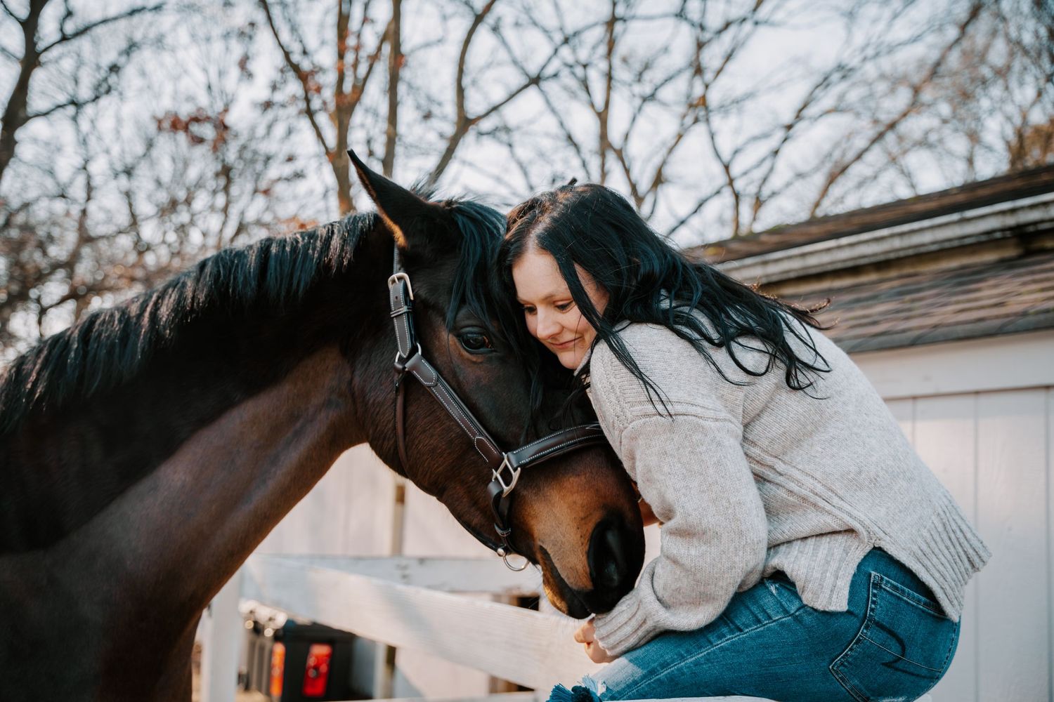 Woman in gray sweater hugging a dark bay horse outdoors near a white barn in winter.