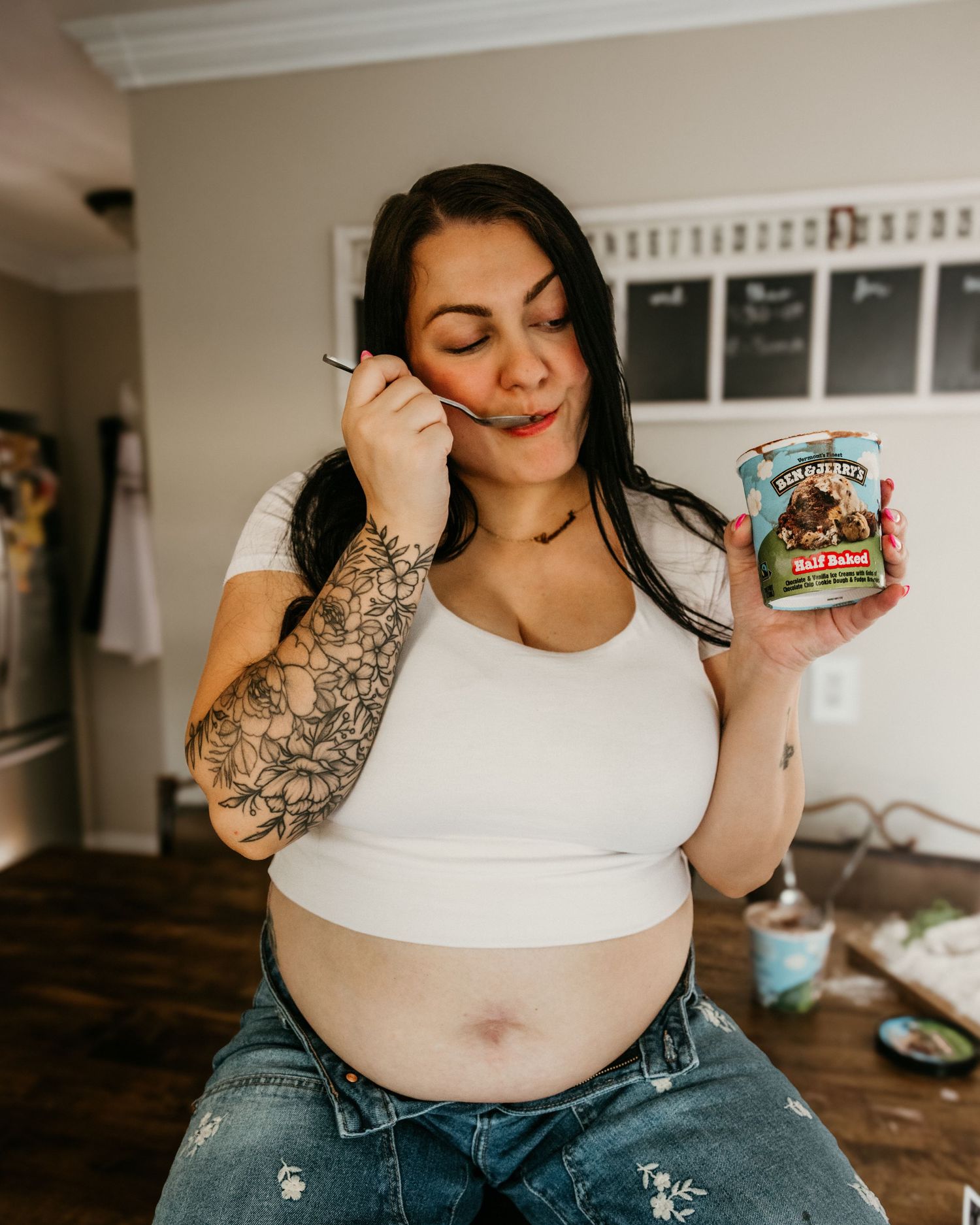 Tattooed woman enjoying Ben & Jerry's Half Baked ice cream directly from the pint in a cozy kitchen.