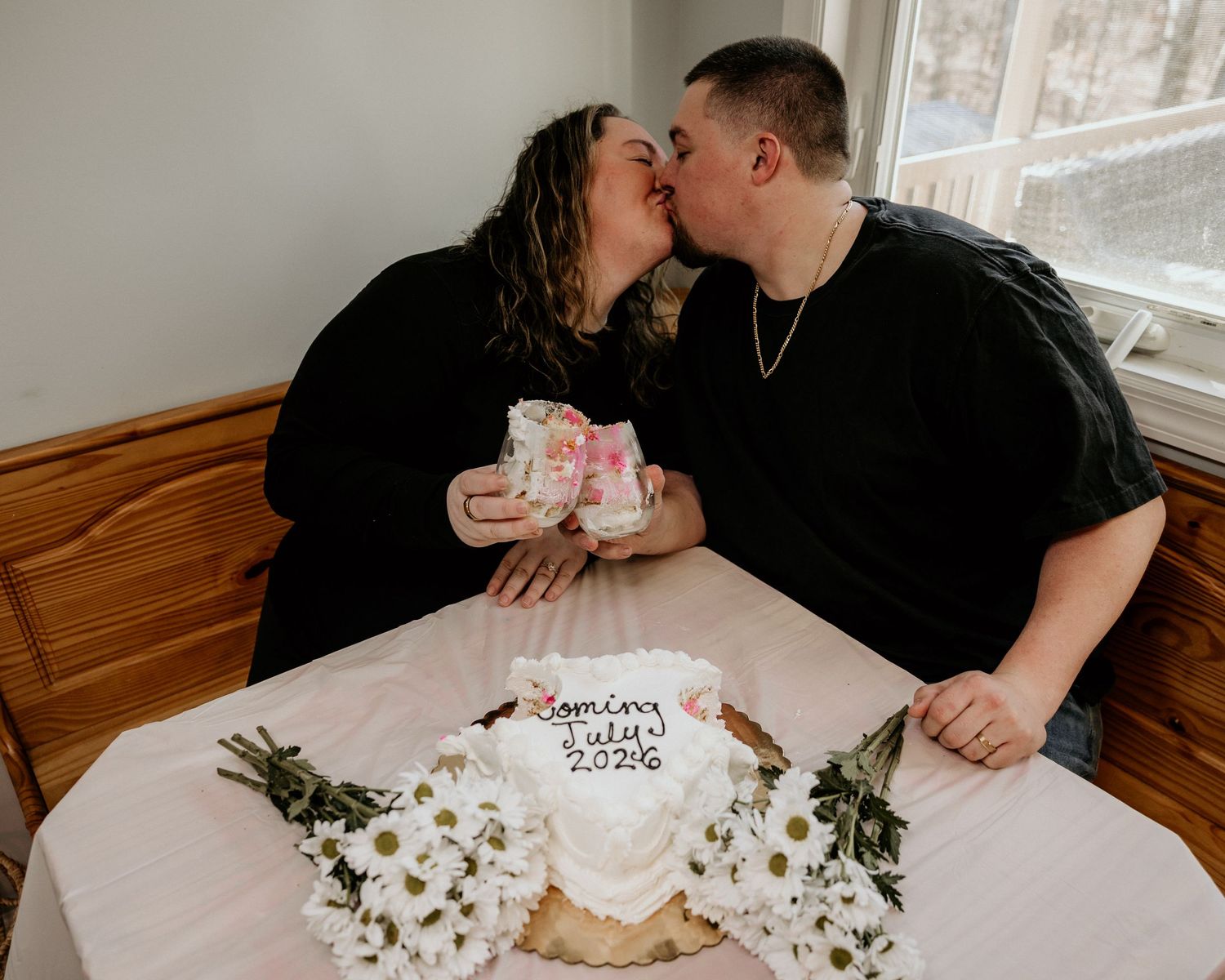 Couple kissing at table with gender reveal cake reading coming July 2026, surrounded by white daisies and pink floral mugs.