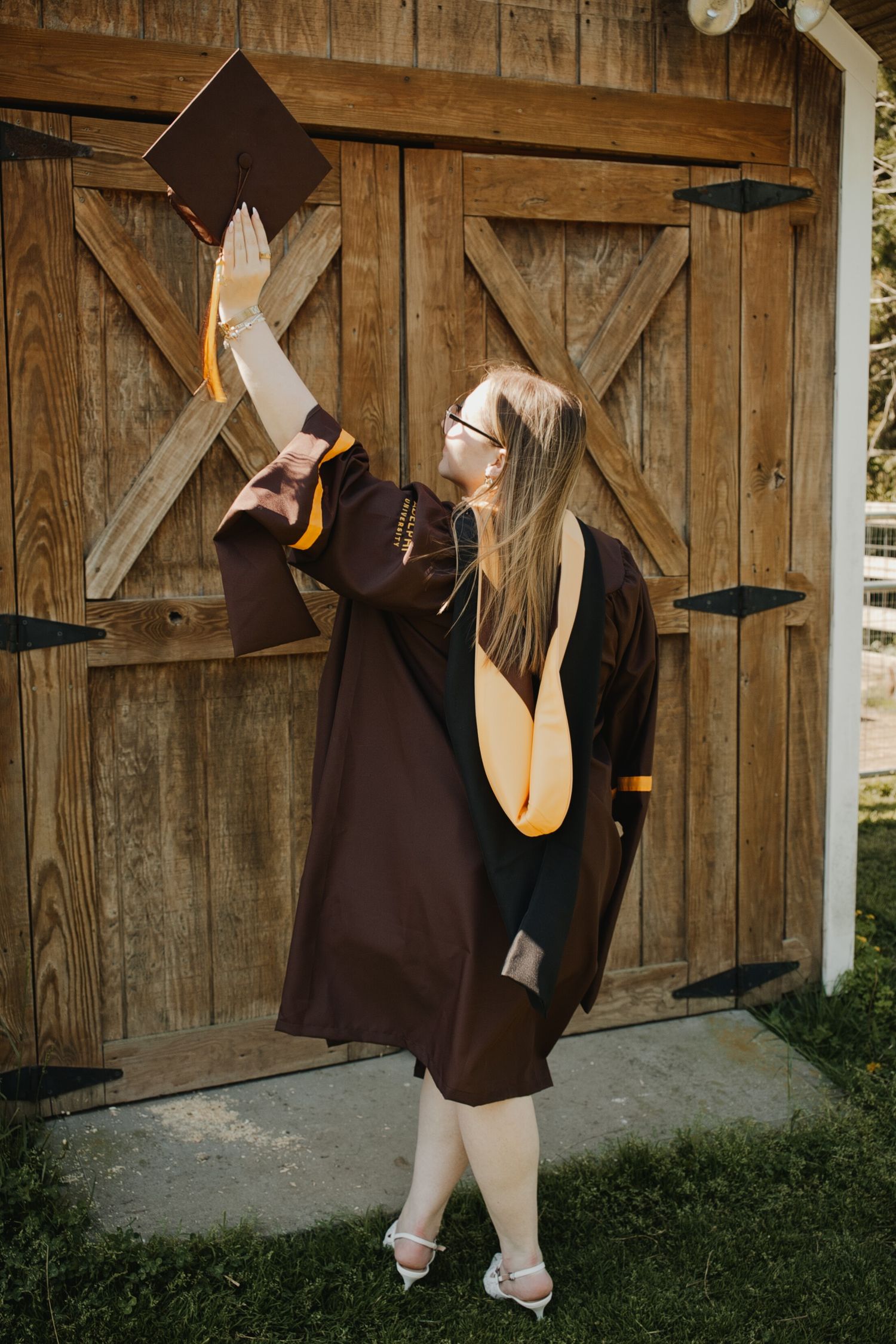 Graduate in brown cap and gown raises her mortarboard triumphantly in front of rustic wooden barn doors.