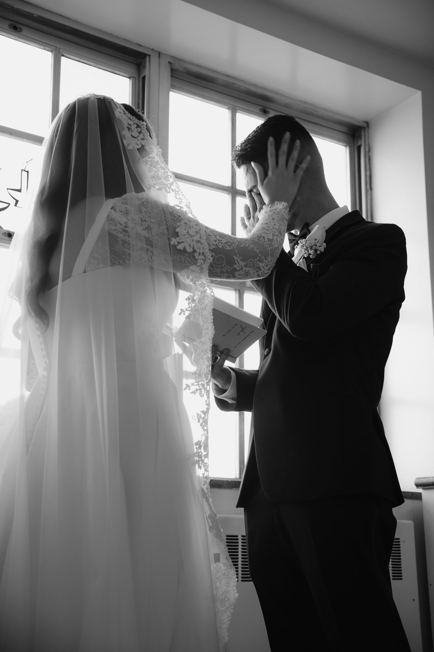 Black and white photo of emotional groom reading vows while bride in lace wedding dress and veil stands by window.