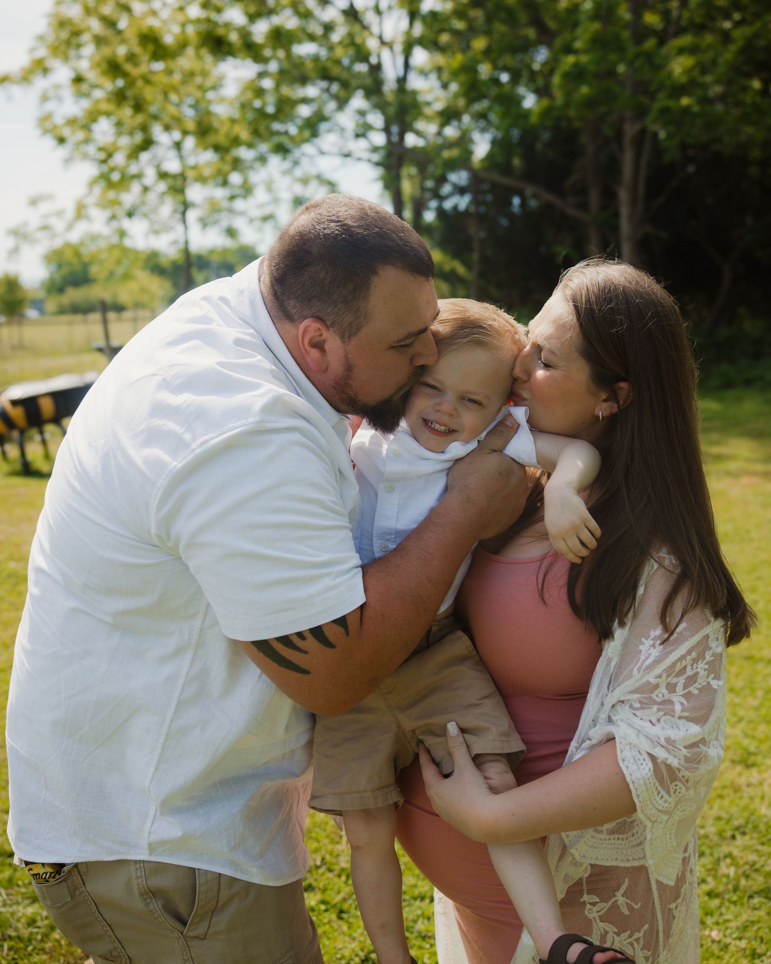 A couple kisses their toddler on the cheeks outdoors in a sunny green park during a family photo session.