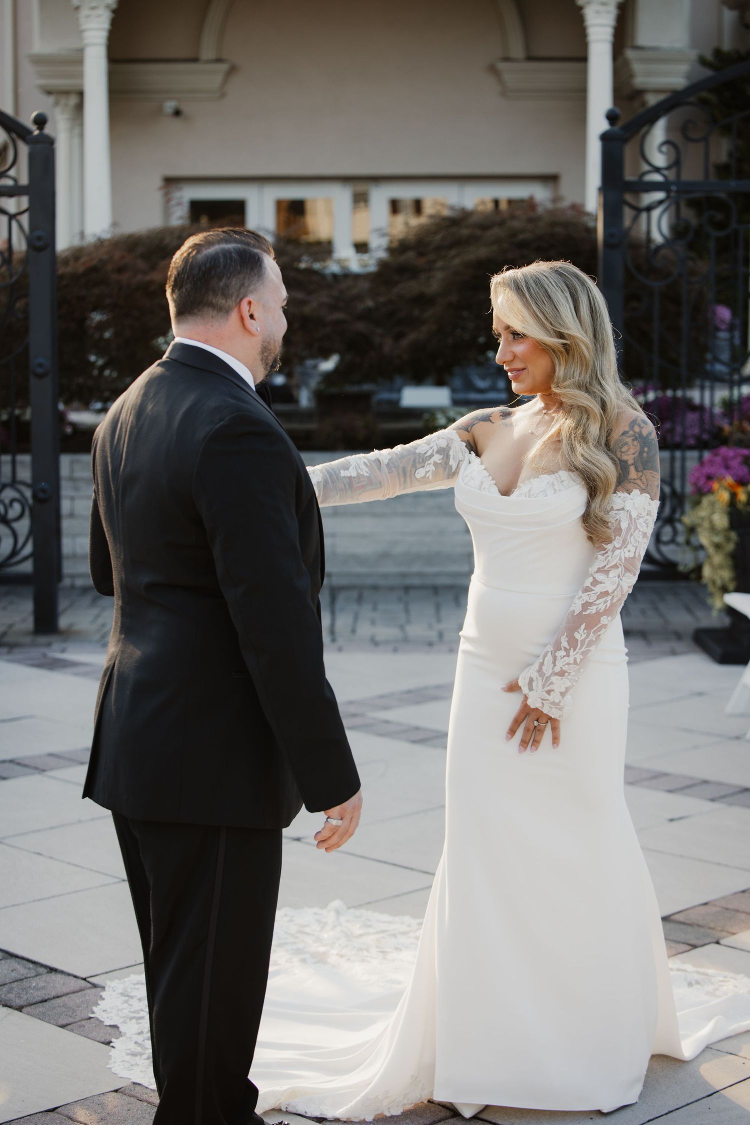 Bride in lace wedding gown touches groom's shoulder during first look outside elegant venue.