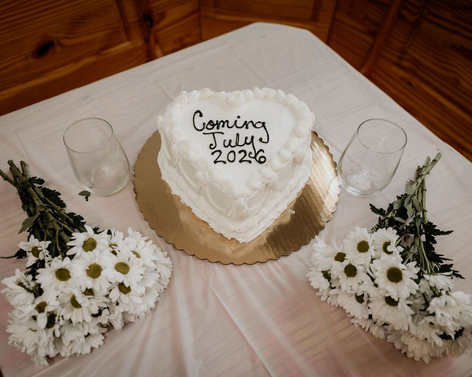 Heart-shaped white cake reading Coming July 2026 on gold stand, flanked by white daisy bouquets and champagne glasses.