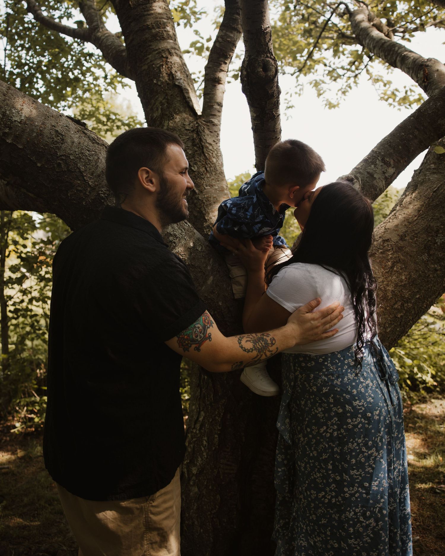 A couple shares a tender moment kissing their baby near a large tree in a lush outdoor setting.