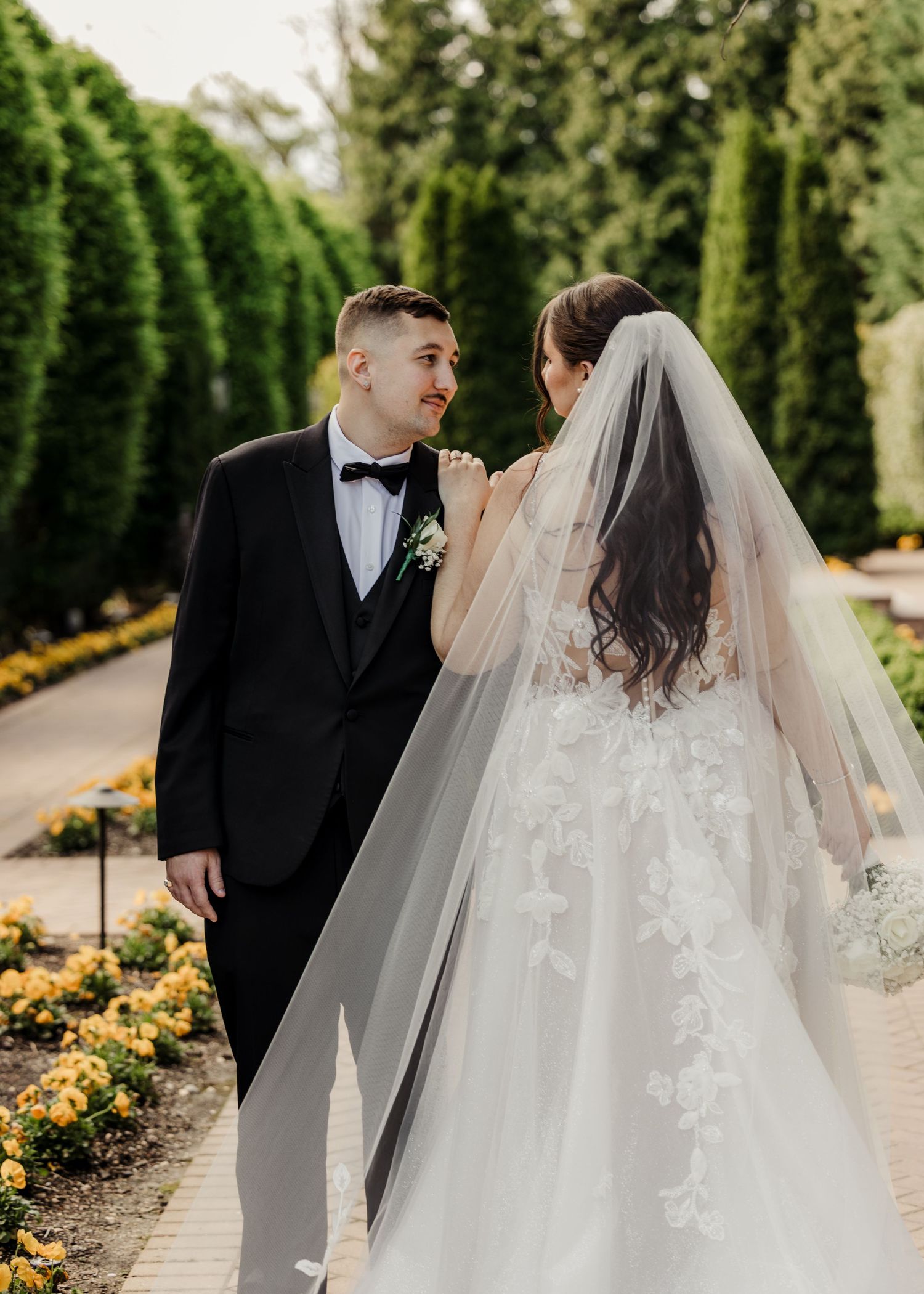 A bride and groom share a romantic moment in a garden, she in a lace gown with veil, he in a black tuxedo.
