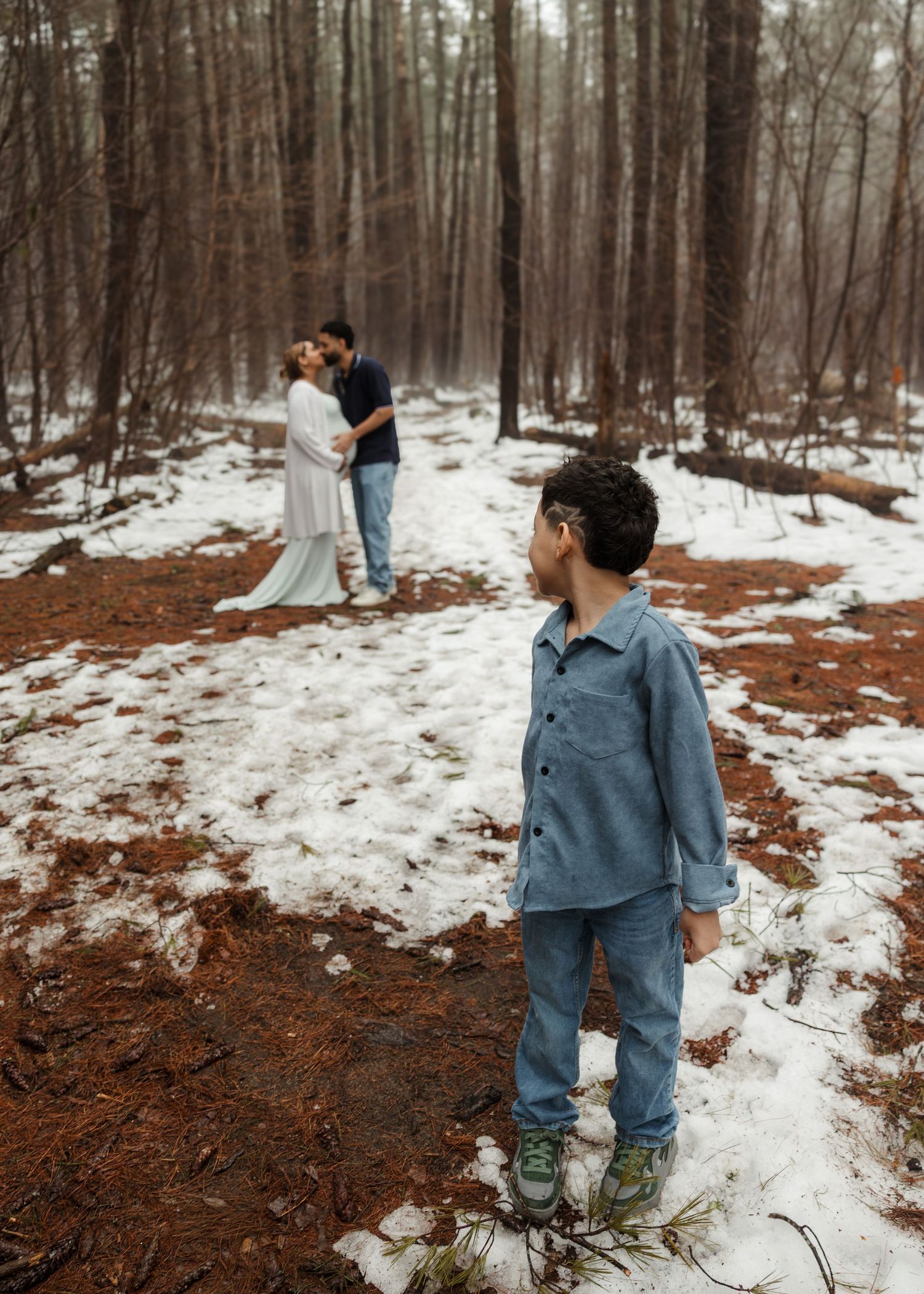 A young boy stands in a snowy forest while a couple kisses in the background during a winter maternity photoshoot.