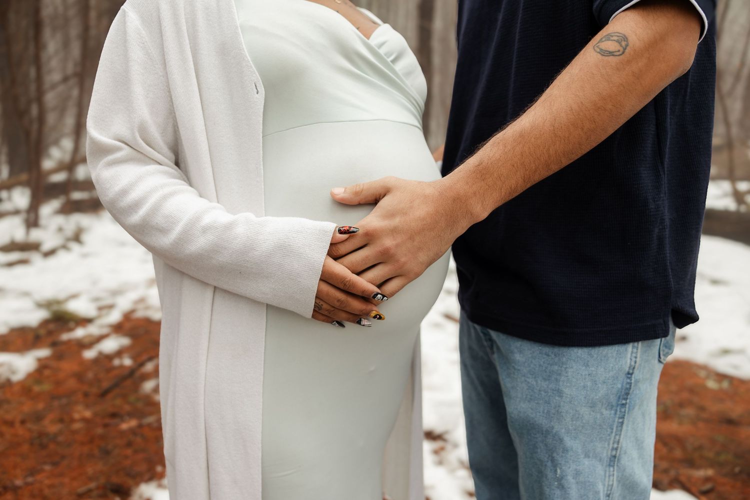 Pregnant woman in white dress with partner touching her baby bump in snowy forest setting.