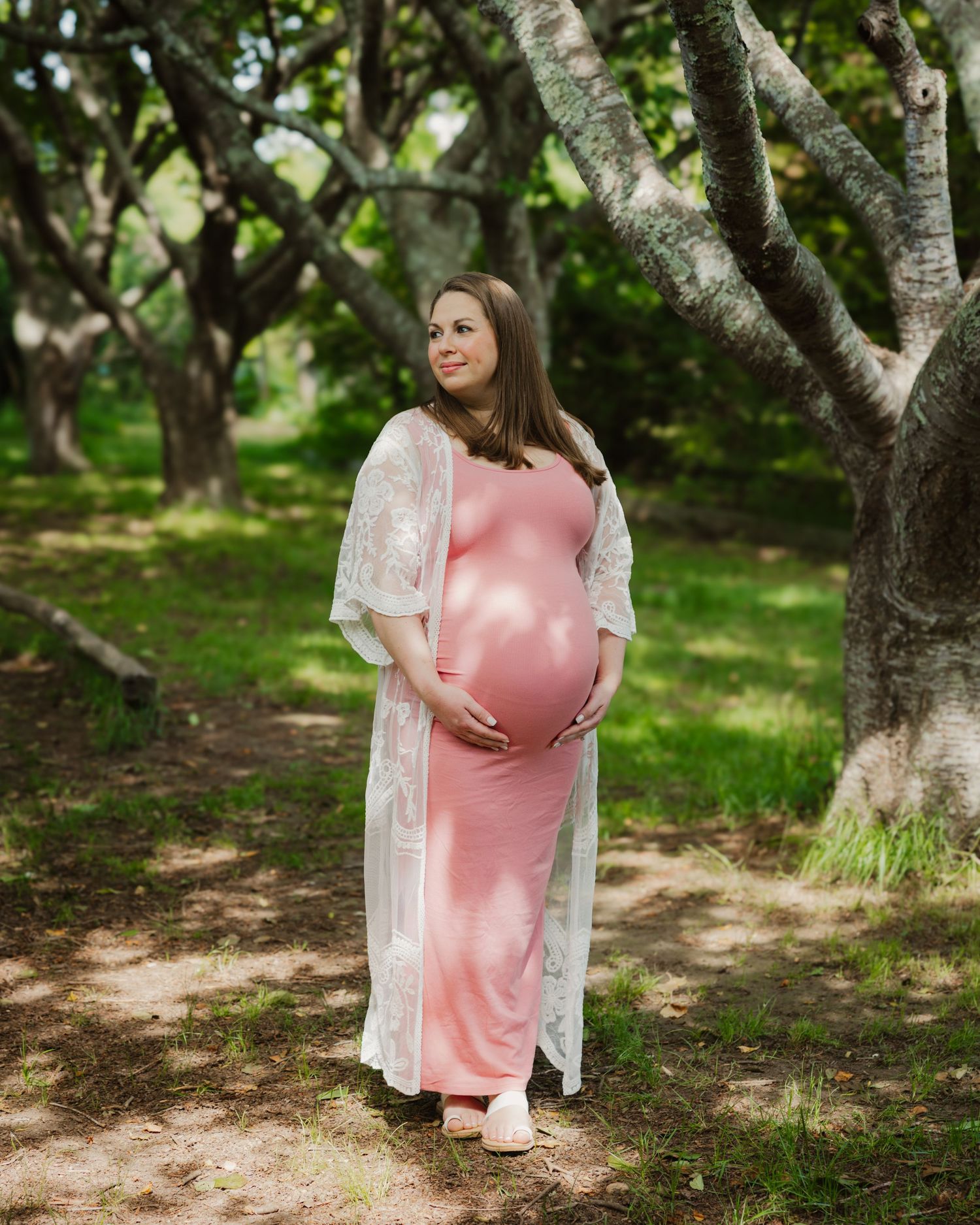 Pregnant woman in pink maxi dress and white lace kimono cradling baby bump outdoors among trees.